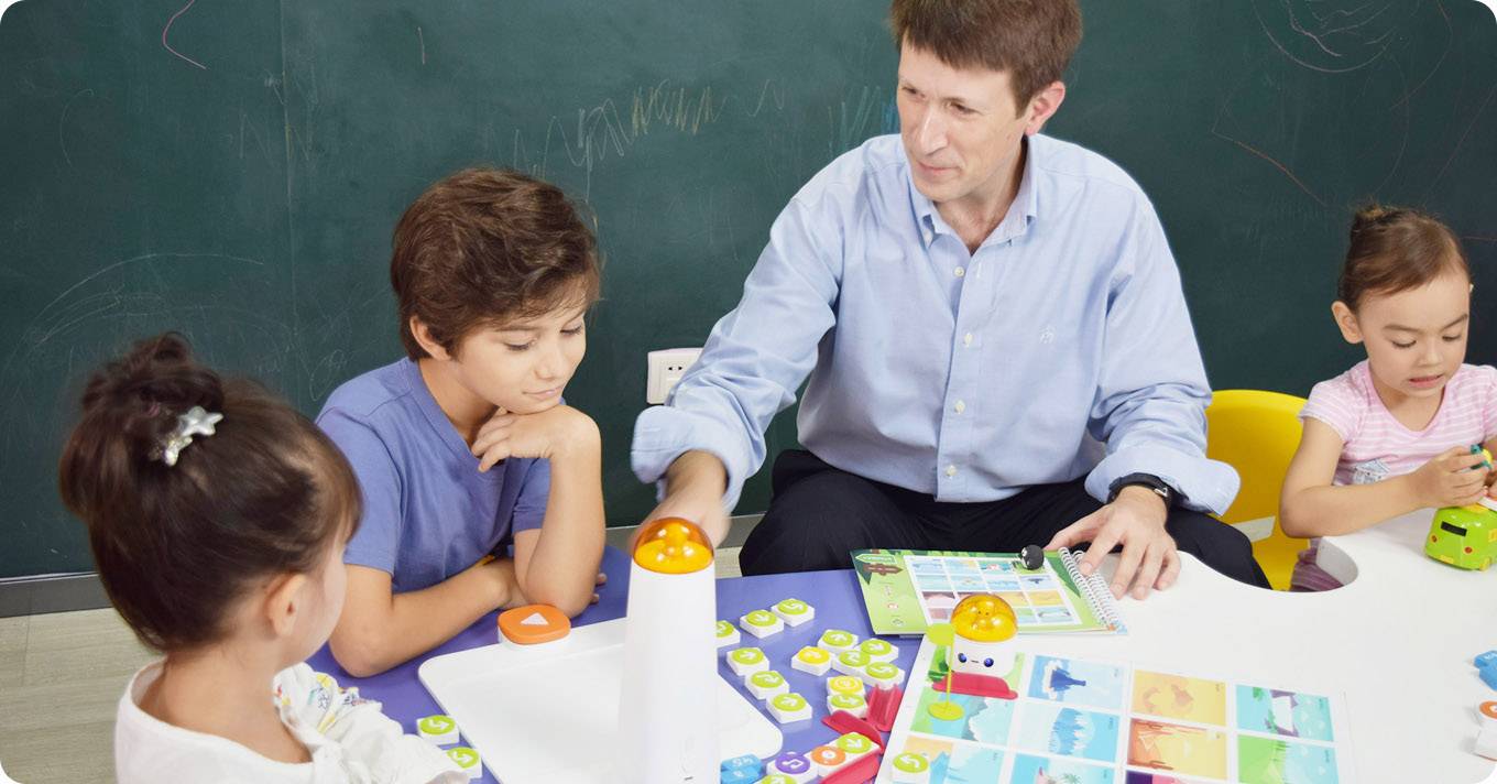 Ein Mann sitzt mit drei Kindern an einem Tisch und spielt ein pädagogisches Brettspiel. Eine grüne Tafel ist im Hintergrund.