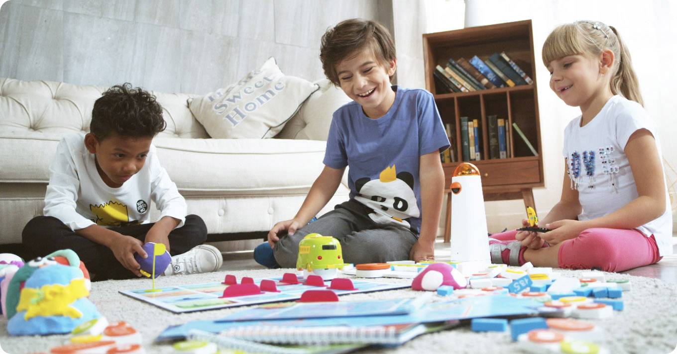 Drei Kinder sitzen am Boden und spielen ein Lernspiel mit bunten Spielzeugen und Karten. Im Hintergrund steht ein Bücherregal.