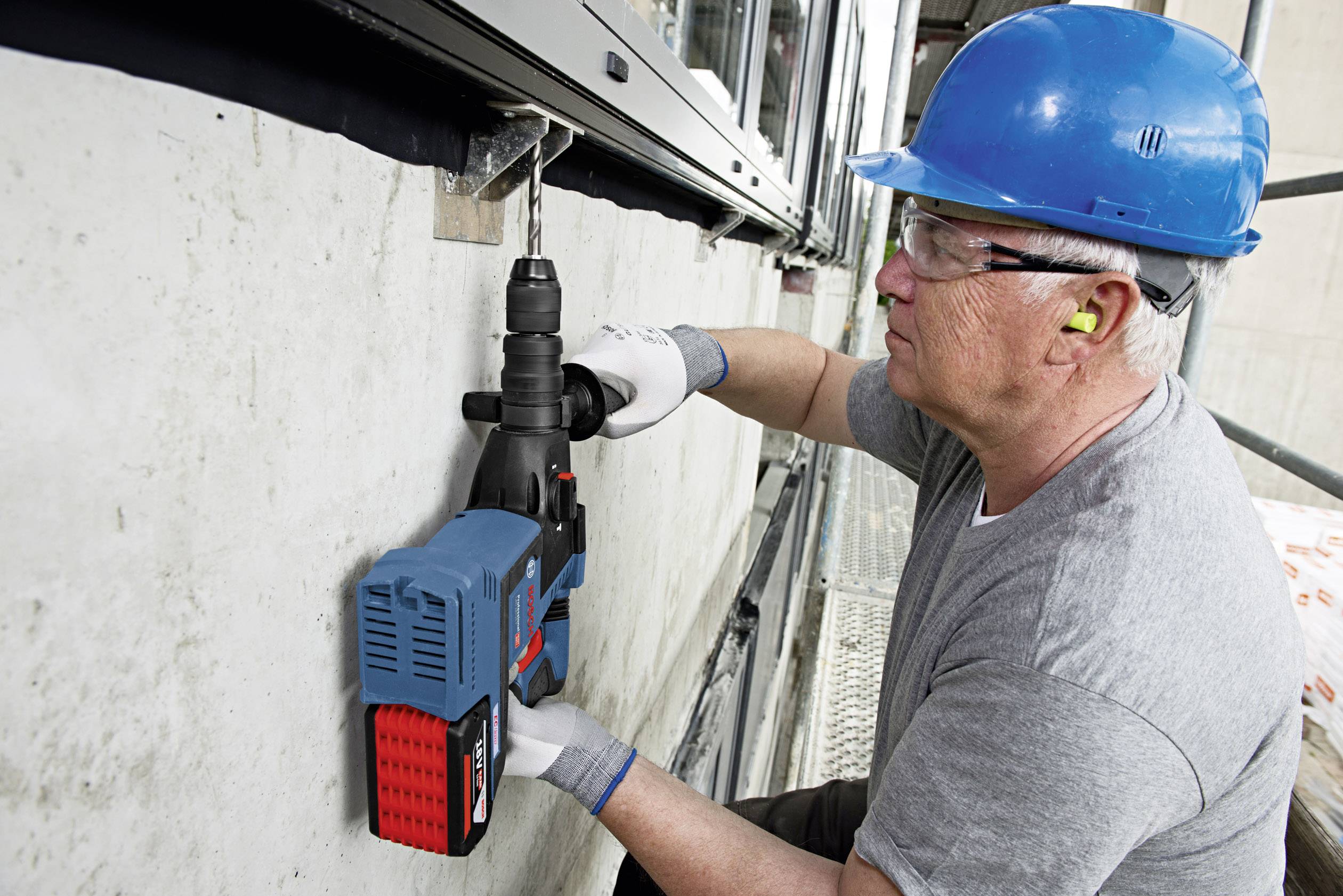 Eine Person mit Schutzbrille und blauem Helm bedient eine Elektrowerkzeugbohrmaschine, um Löcher in eine Betonwand zu bohren.