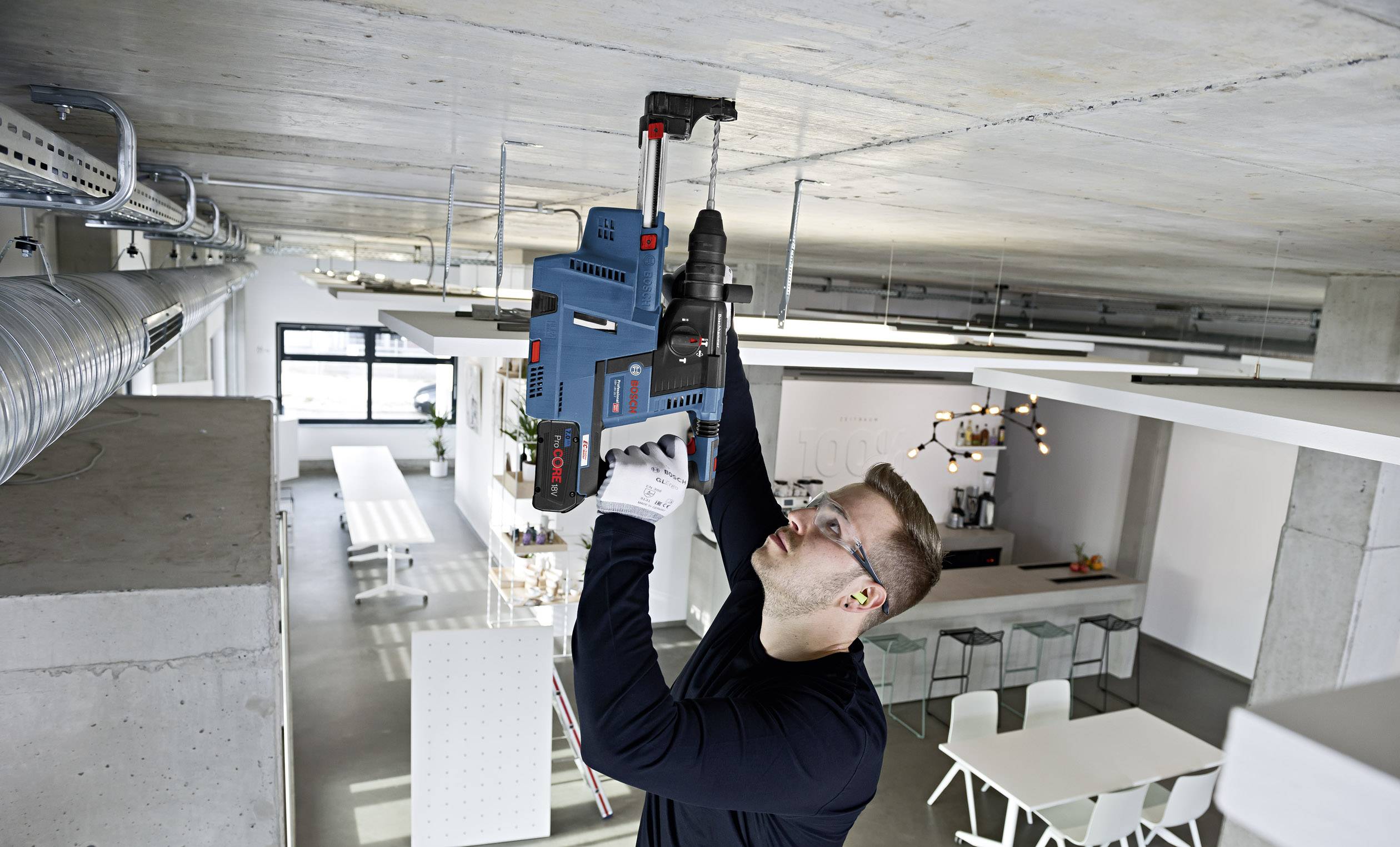Ein Mann bohrt mit einem großen Elektrobohrer in eine Decke in einem modernen Bürogebäude. Er trägt Schutzbrille und Handschuhe.