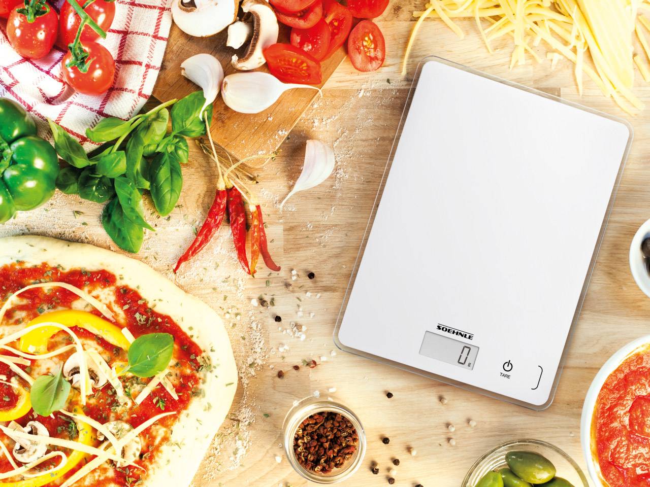 Digitale Küchenwaage auf Holztisch, umgeben von frischen Pizzazutaten wie Teig, Gemüse, Kräuter und Gewürze. Display zeigt '0'.