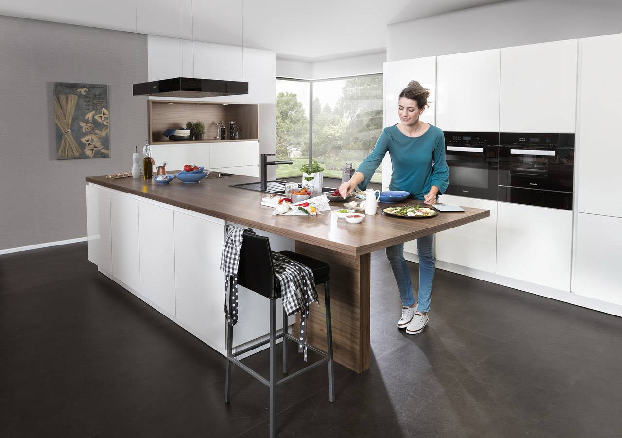 Eine Frau bereitet in einer modernen, hellen Küche Essen zu. Auf der Arbeitsplatte stehen Gewürze und Lebensmittel, im Hintergrund ist ein großes Fenster.