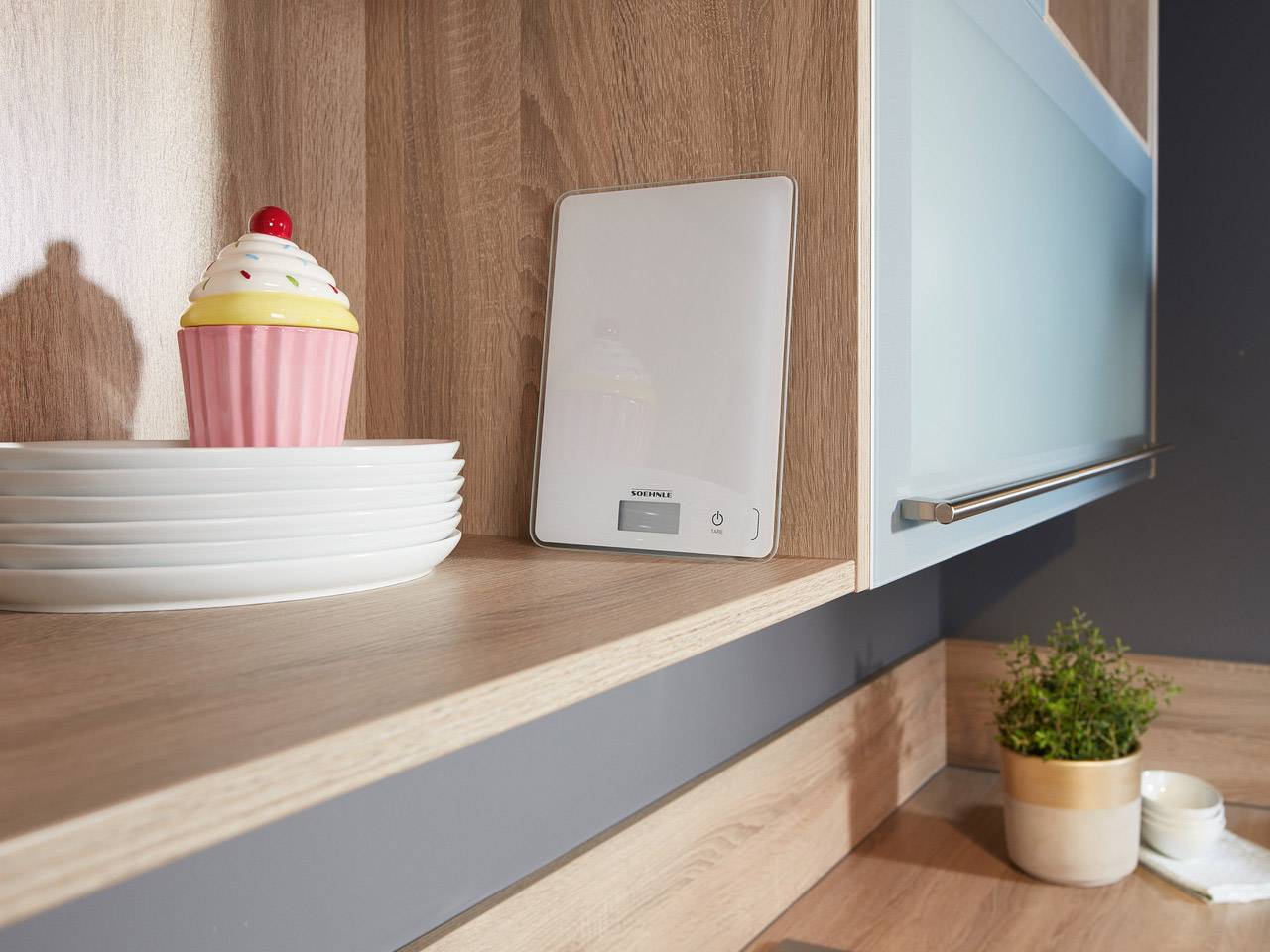 Küchenszene mit digitaler Waage auf Holzregal, daneben ein kleiner Blumentopf und ein Stapel Teller mit dekorativem Muffin-Design.