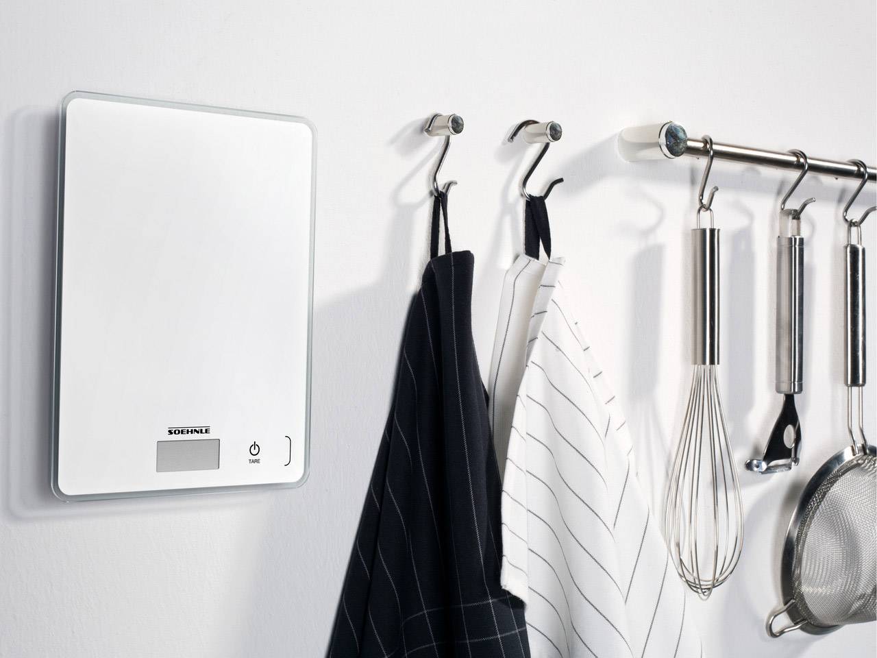 Digitale Küchenwaage an der Wand, daneben hängen Küchentücher und -utensilien an Haken.