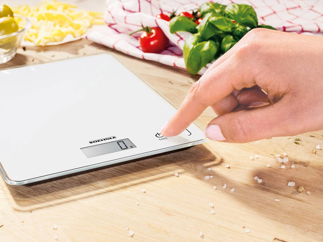 Eine Hand drückt den Einschaltknopf einer digitalen Küchenwaage auf einem Holztisch. Im Hintergrund sind Tomaten und Gewürze.