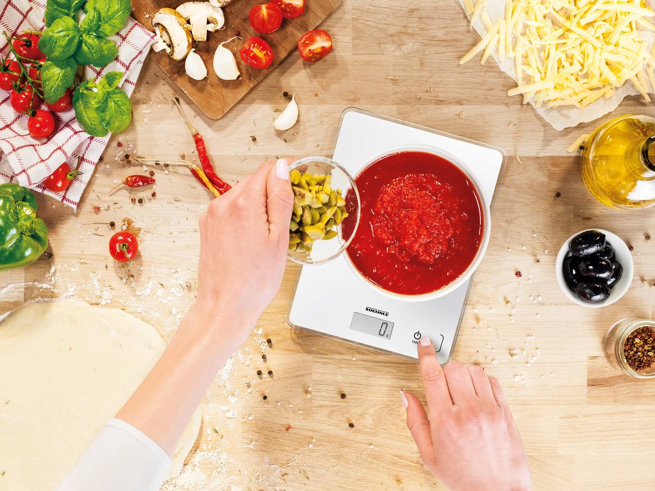 Person wiegt gehackte Oliven auf einer digitalen Küchenwaage neben einer Schüssel mit Tomatensauce, umgeben von Pizzazutaten.
