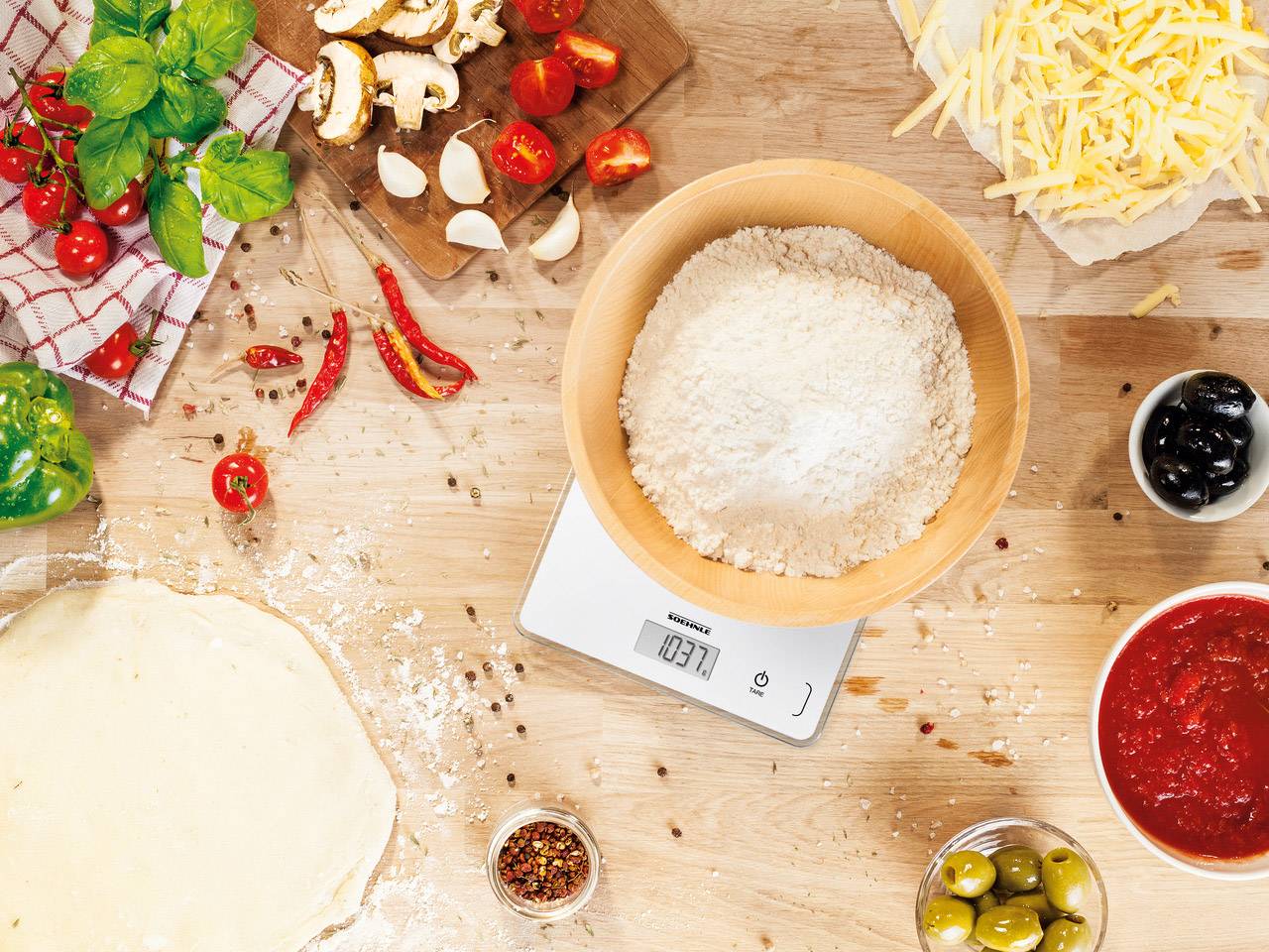 Backzutaten auf Holzbrett mit digitaler Waage: Mehl in Schüssel, Tomatensauce, geriebener Käse, Tomaten, Pilze, Oliven. Pizza-Zubereitung.