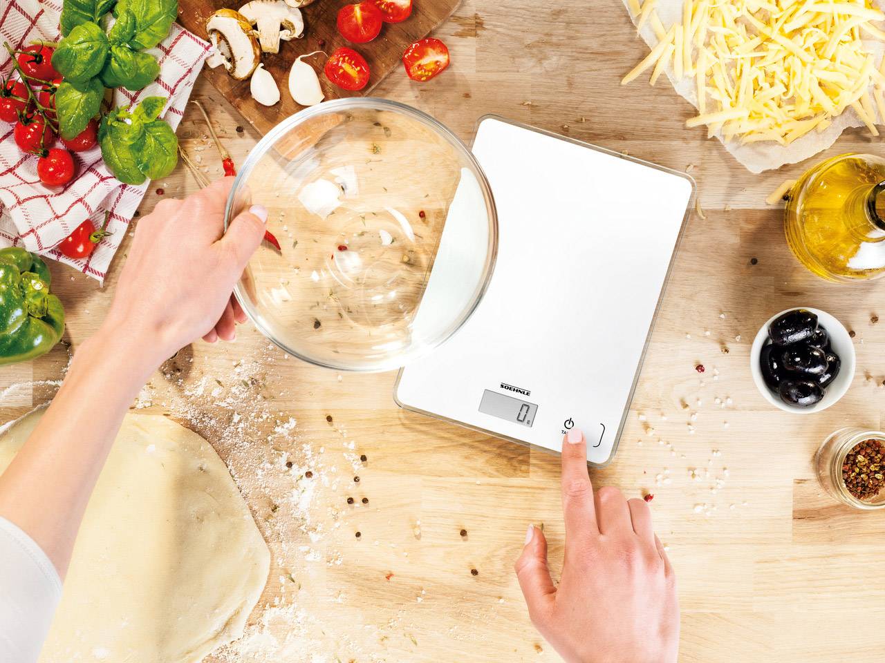 Eine Person wiegt eine Glasschüssel auf einer digitalen Küchenwaage, umgeben von Zutaten wie Basilikum, Tomaten, Oliven und Käse.