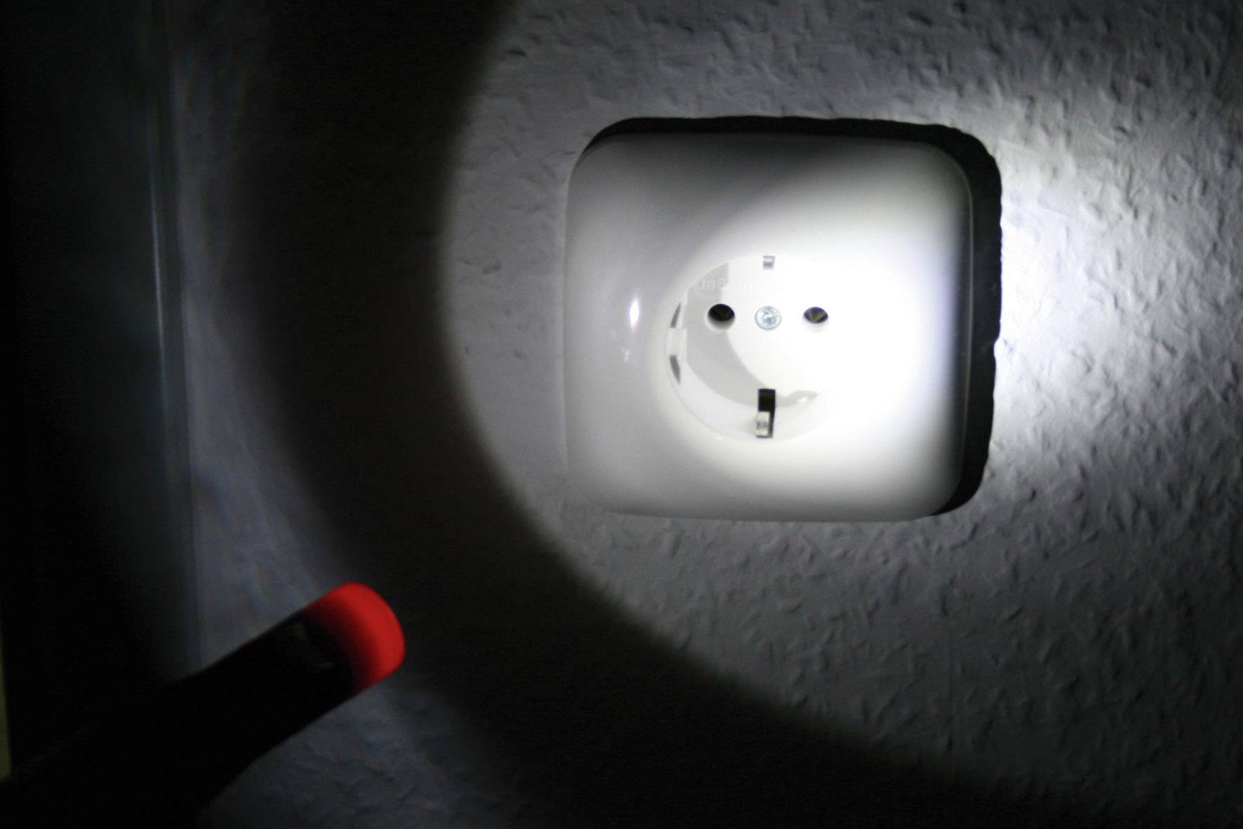 Eine Person beleuchtet in dunkler Umgebung mit einer Taschenlampe eine Wandsteckdose.