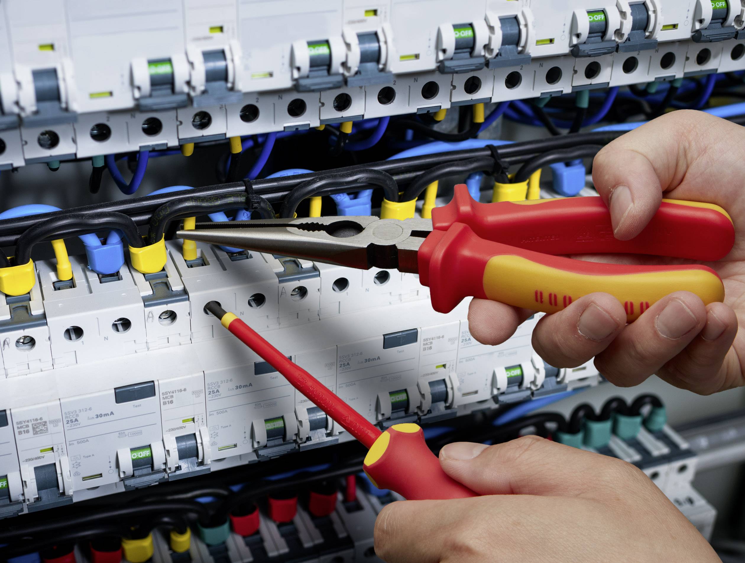 Eine Person repariert eine elektrische Schalttafel mit einer Zange und einem Schraubendreher. Kabel und Sicherungen sind sichtbar.