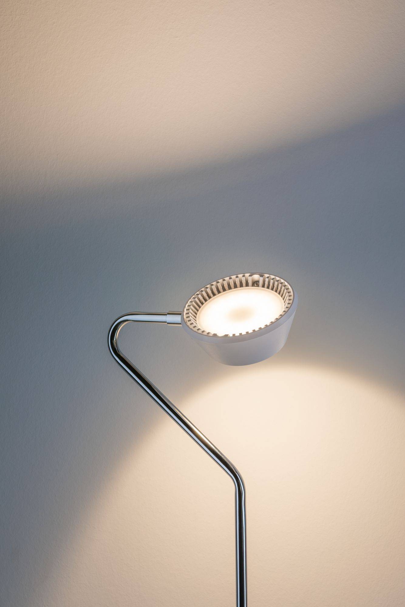 Eine moderne, minimalistische Stehlampe leuchtet gegen eine Wand. Die Lampe hat einen metallischen, gebogenen Arm und einen runden Lampenschirm.
