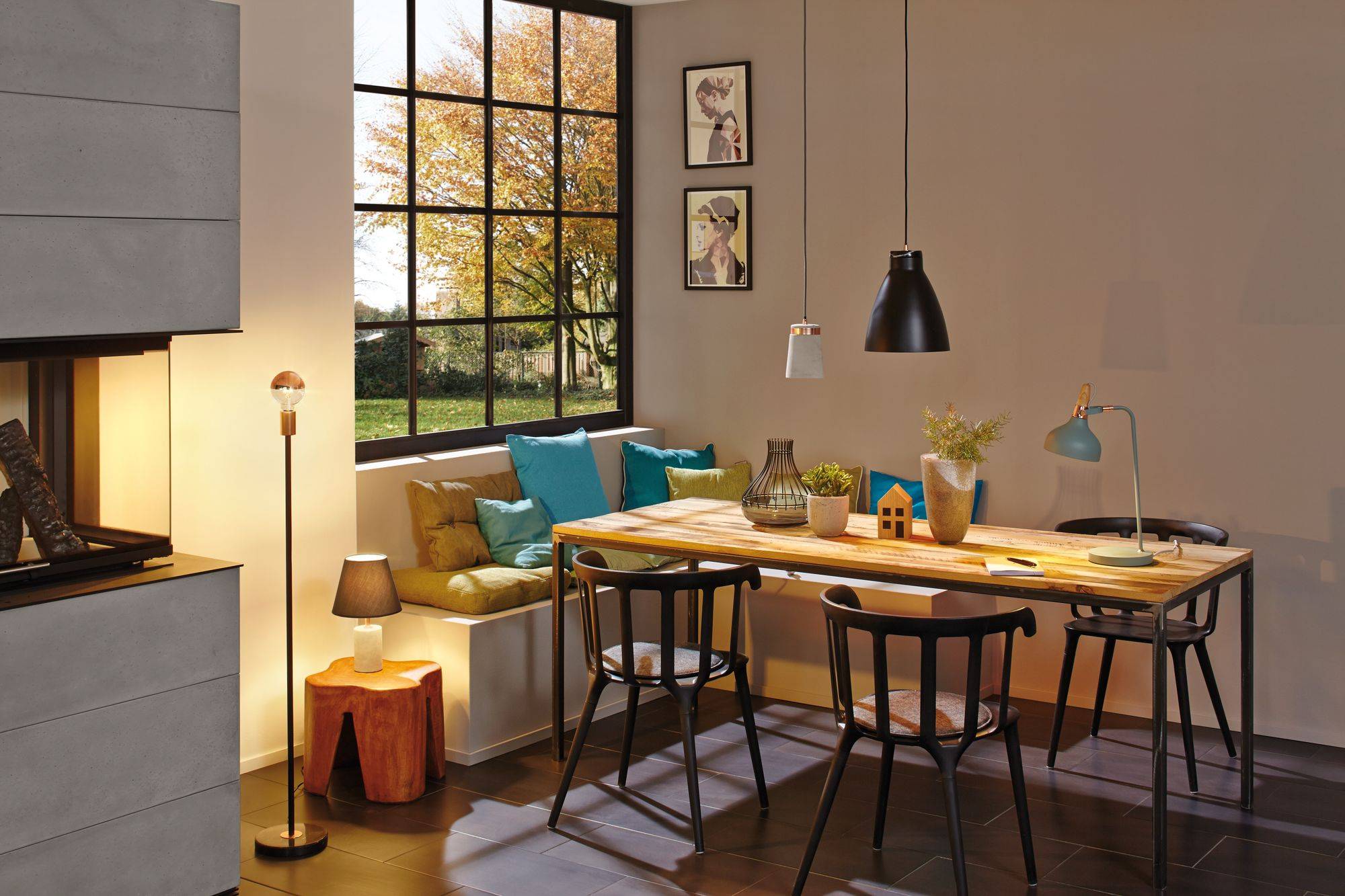 Ein Esszimmer mit großem Holztisch, umgeben von schwarzen Stühlen. Durch das Fenster fällt Sonnenlicht auf bunte Kissen auf einer Bank.