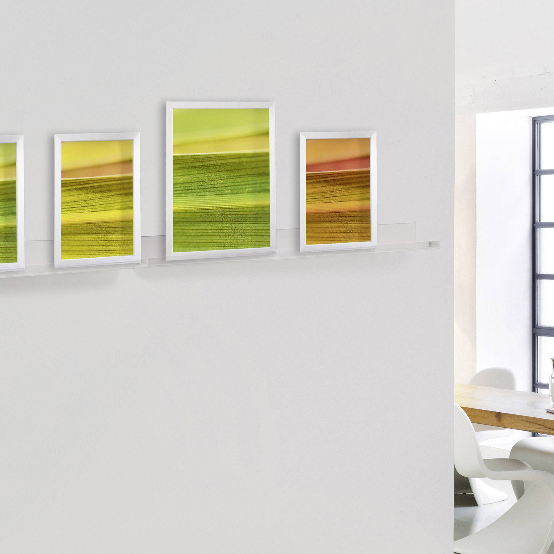 Vier farbige Landschaftsbilder hängen in gleichem Abstand an einer weißen Wand. Rechts davon ein Fenster mit hellem Licht.