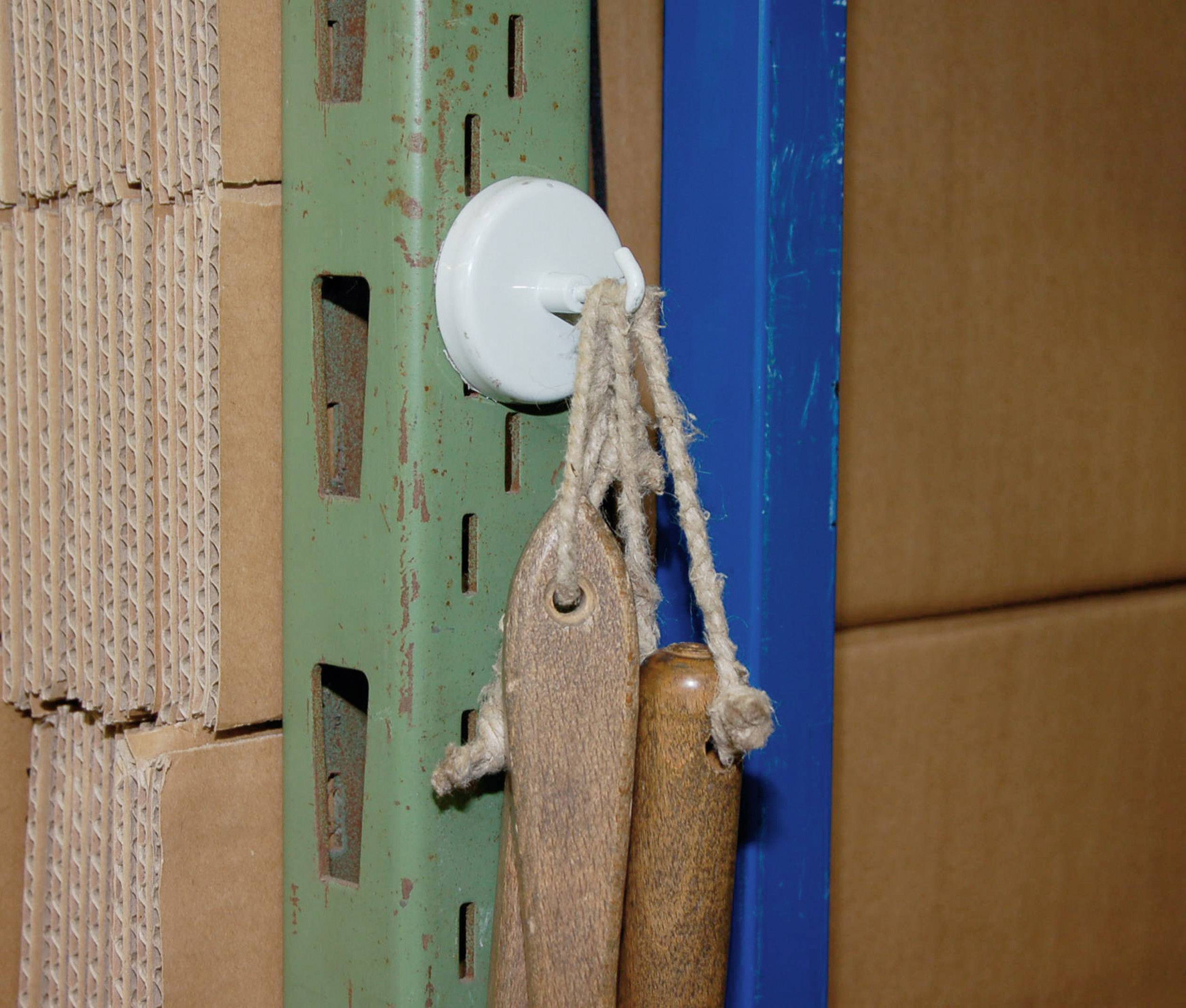 Ein grüner Metallständer hält Werkzeuge an einem weißen Haken; daneben blaue Stange. Kartons im Hintergrund.