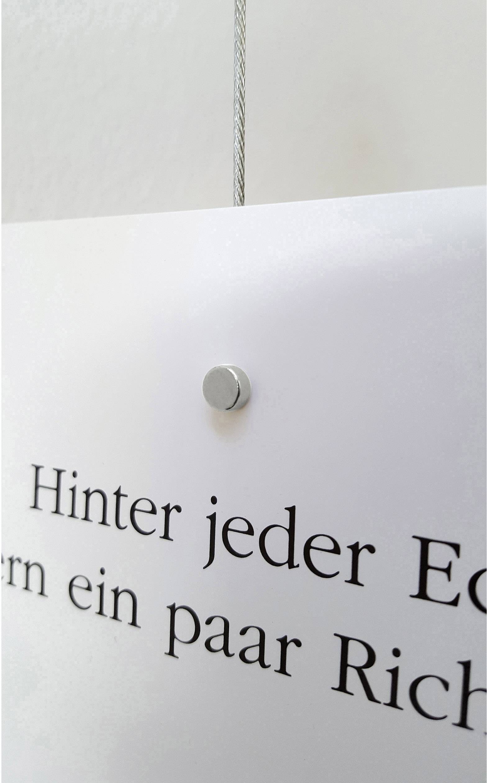 Ein weißes Schild hängt an einem Drahtseil. Es zeigt teilweise den Text 'Hinter jeder Ecke fernern ein paar Rich' in schwarzer Schrift.
