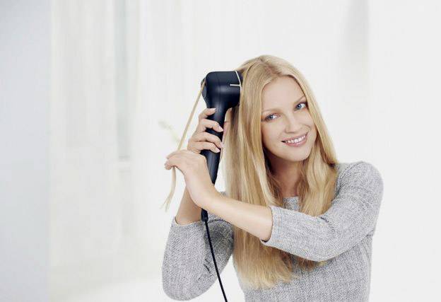 Eine Frau mit langen blonden Haaren verwendet ein schwarzes Haarstyling-Gerät. Sie trägt einen grauen Pullover und lächelt entspannt.