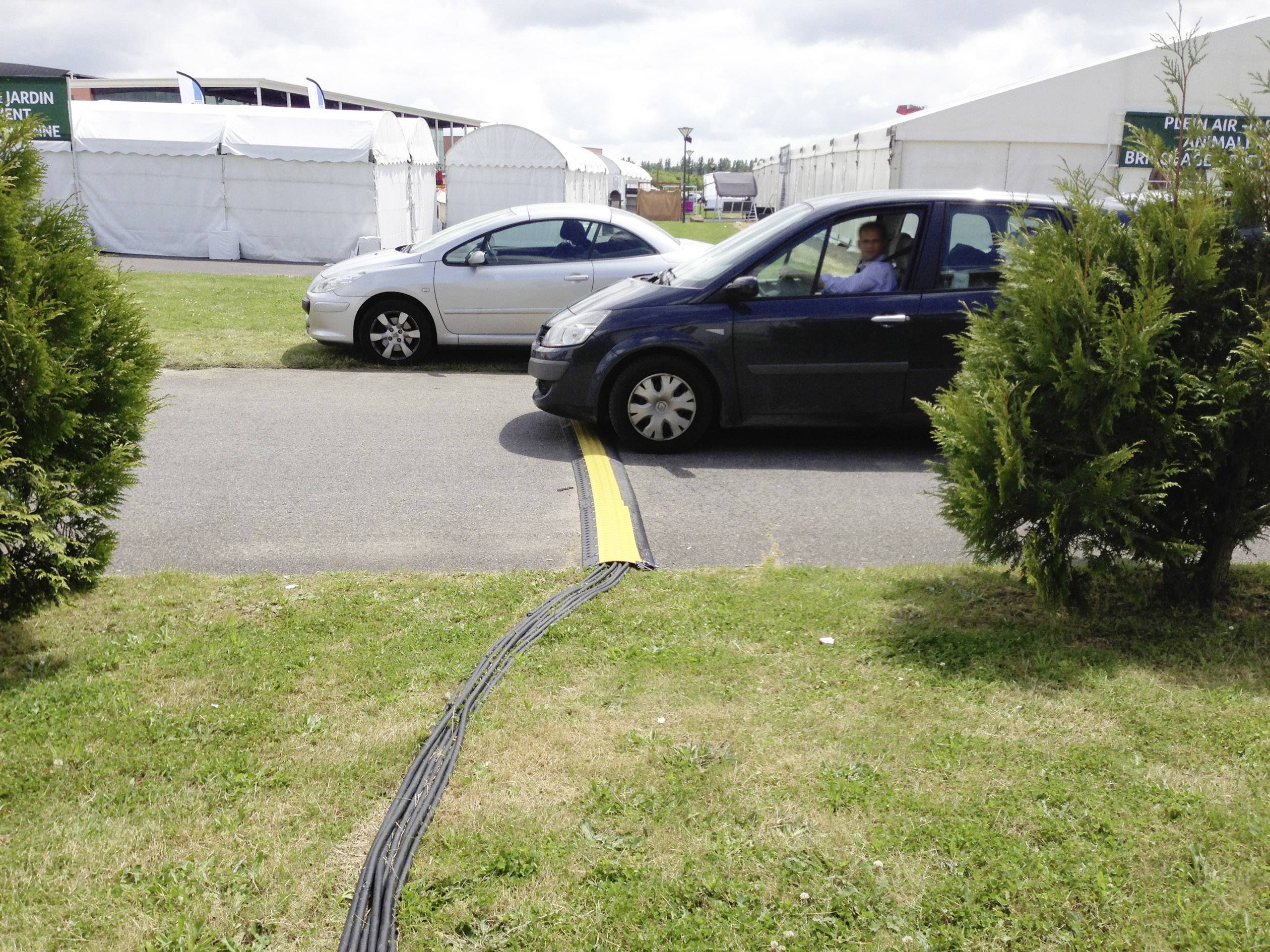 Ein schwarzes Auto fährt vorsichtig über Kabel, die auf einer Straße verlaufen, umgeben von Gras und Zelten im Hintergrund.