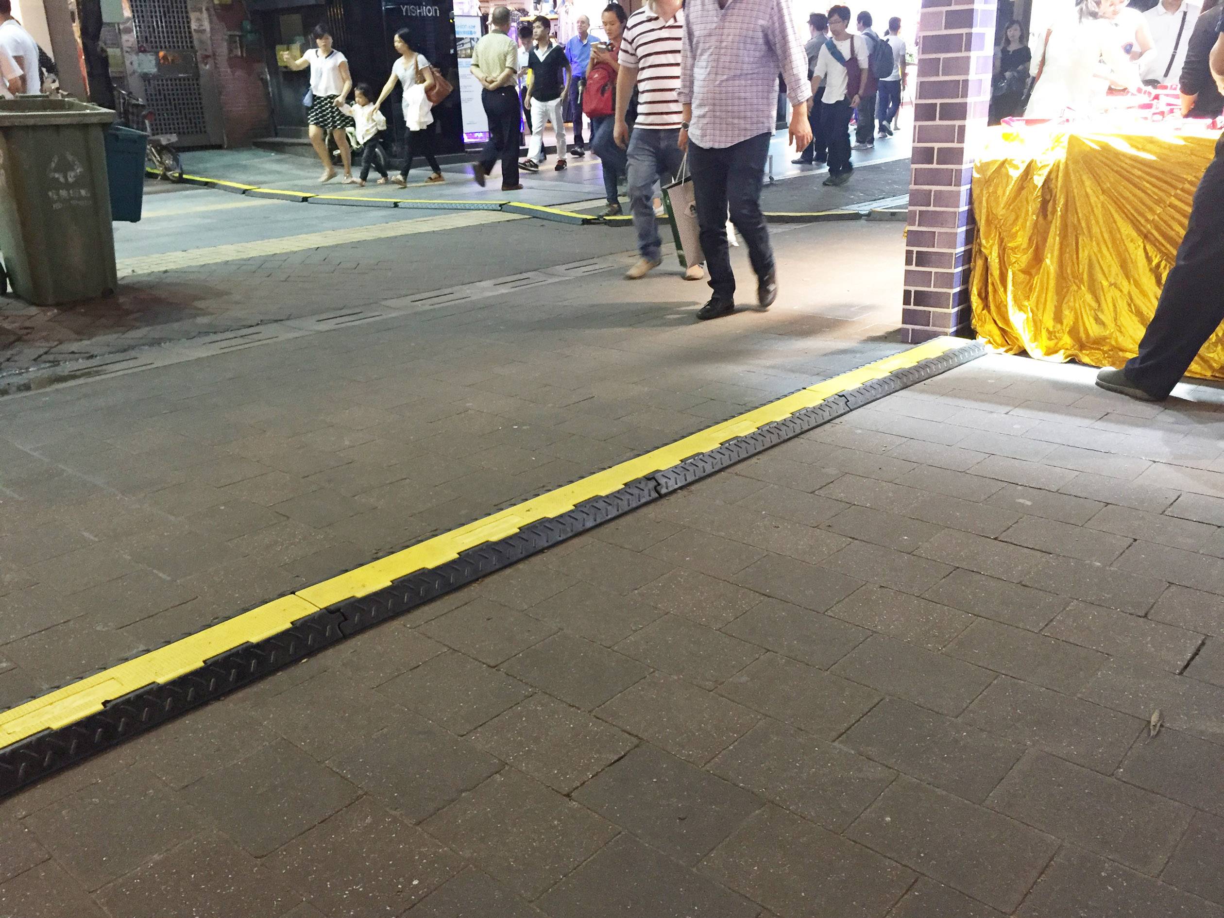 Eine erhöhte Schwelle auf einem Gehweg mit gelben und schwarzen Streifen; Menschen laufen im Hintergrund entlang.