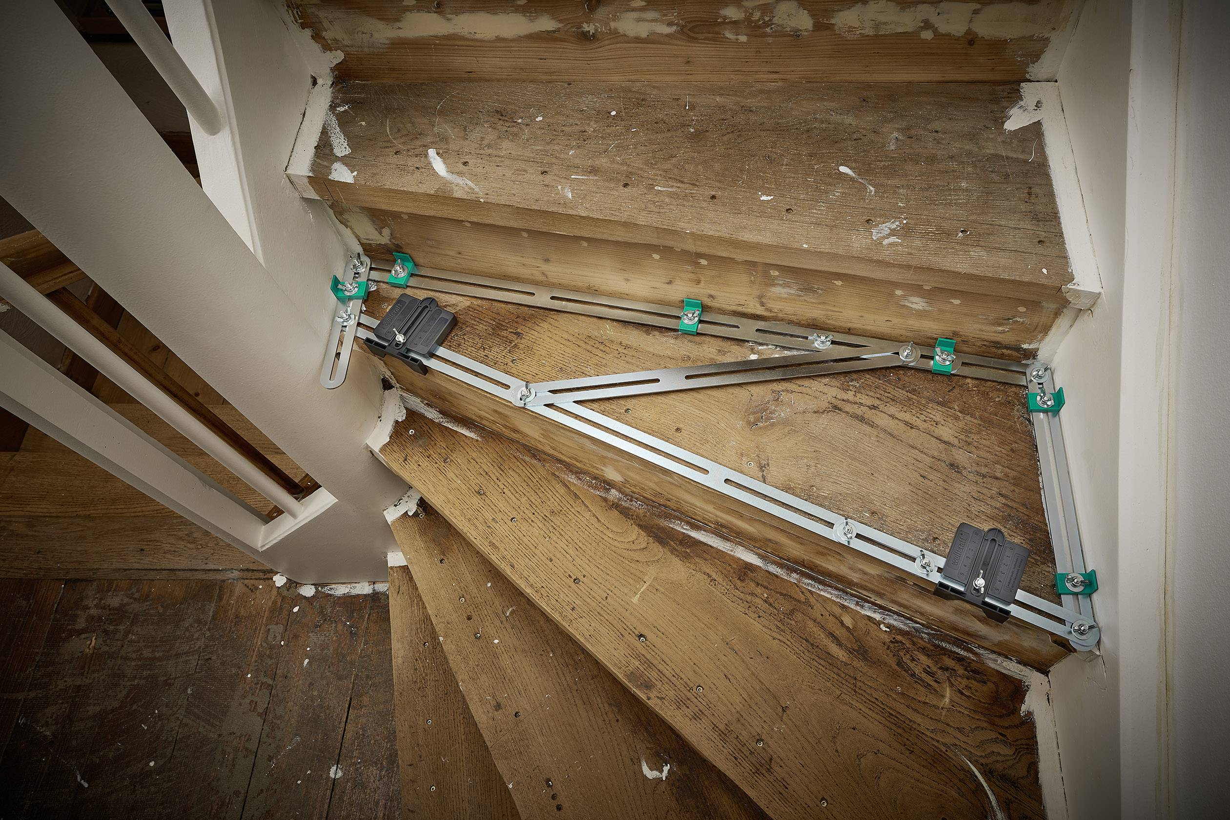 Eine Holztreppe wird mit einem Schablonengerät vermessen. Das Gerät besteht aus Metallstäben mit grünen und schwarzen Klemmen zur Anpassung.