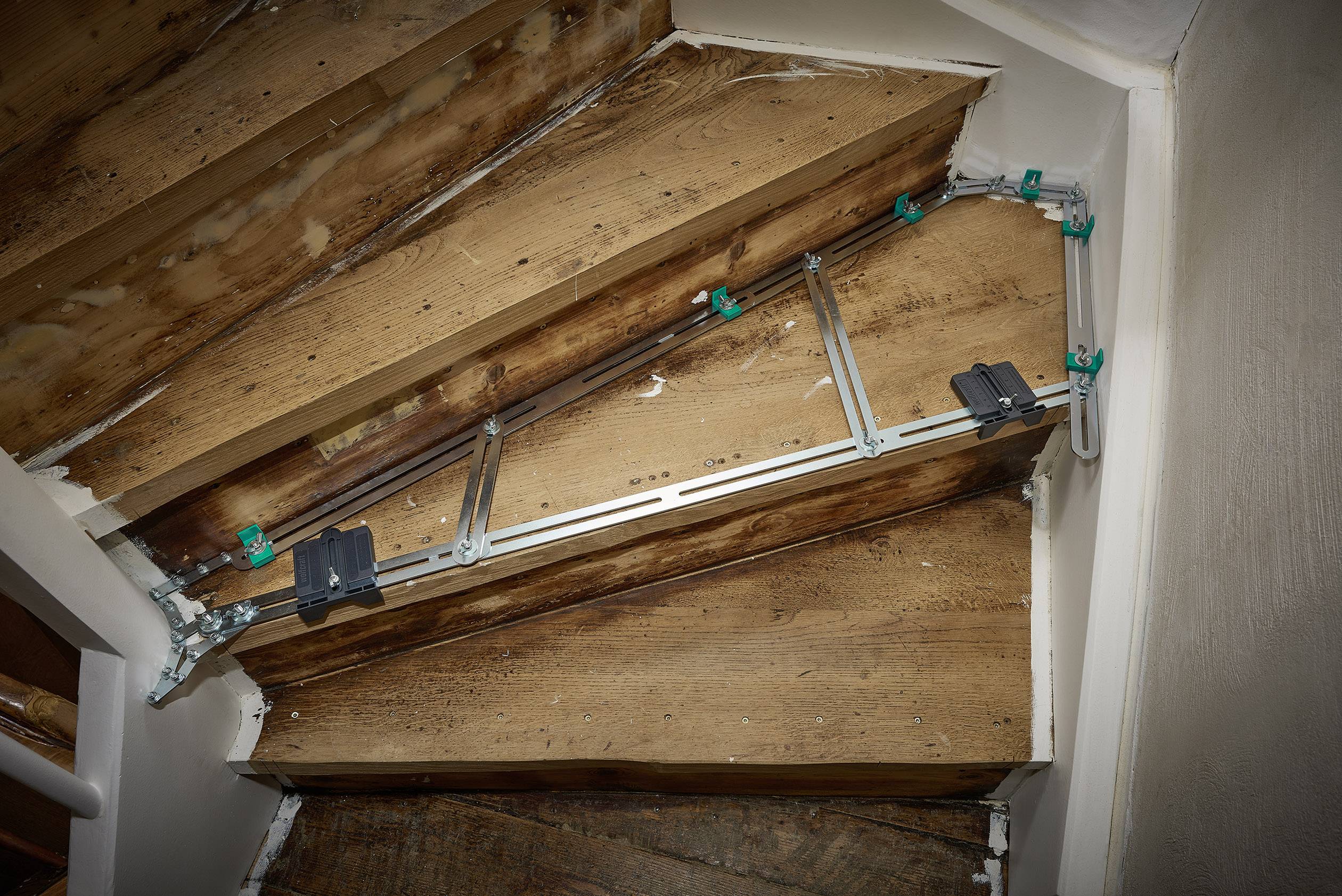 Ein Treppenaufzugssystem ist an einer schrägen Holztreppenkonstruktion montiert und zeigt Metallführungen mit grünen Halterungen.