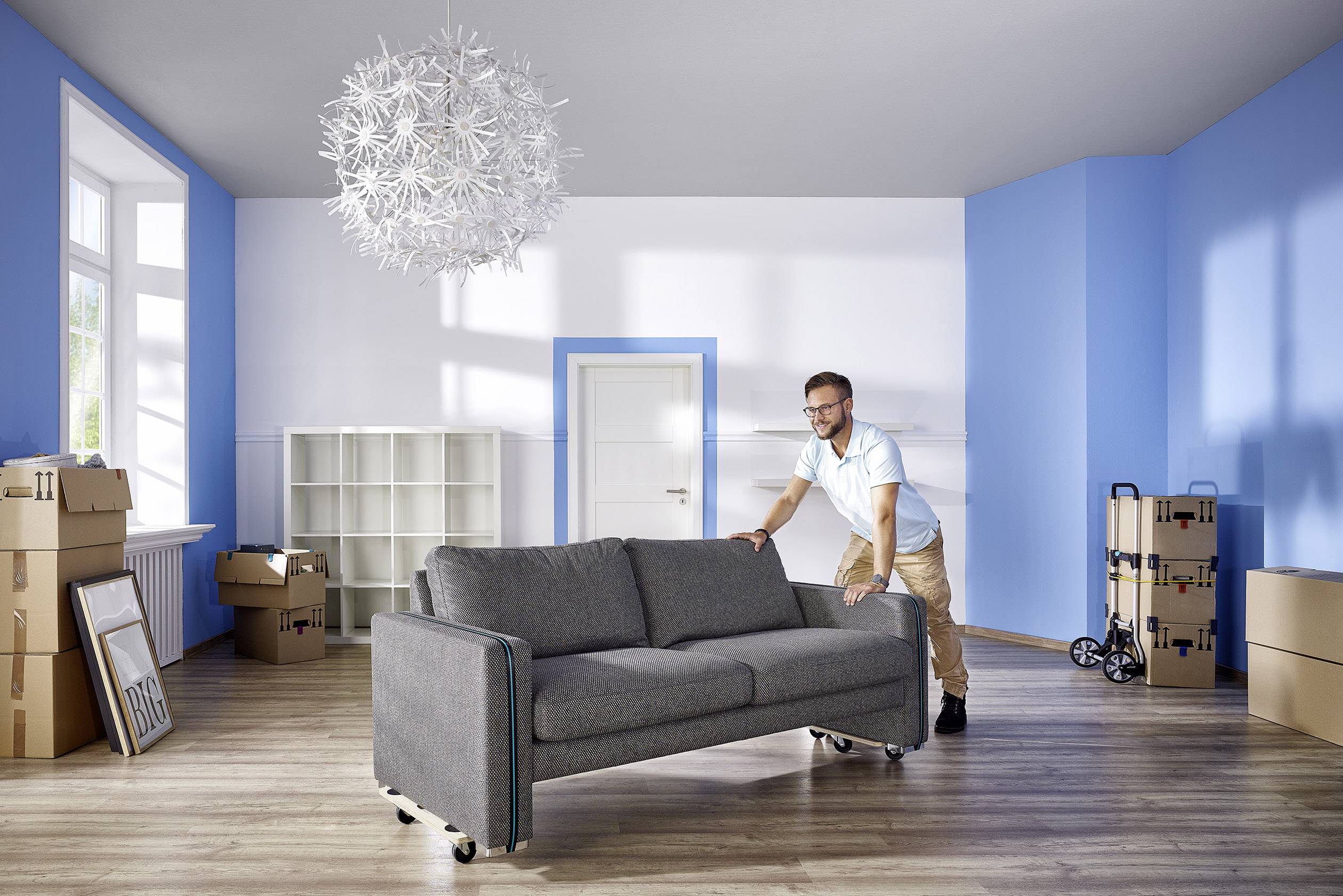 Ein Mann bewegt ein graues Sofa in einem leeren Raum mit blauen Wänden. Umzugskartons stehen im Raum verteilt.