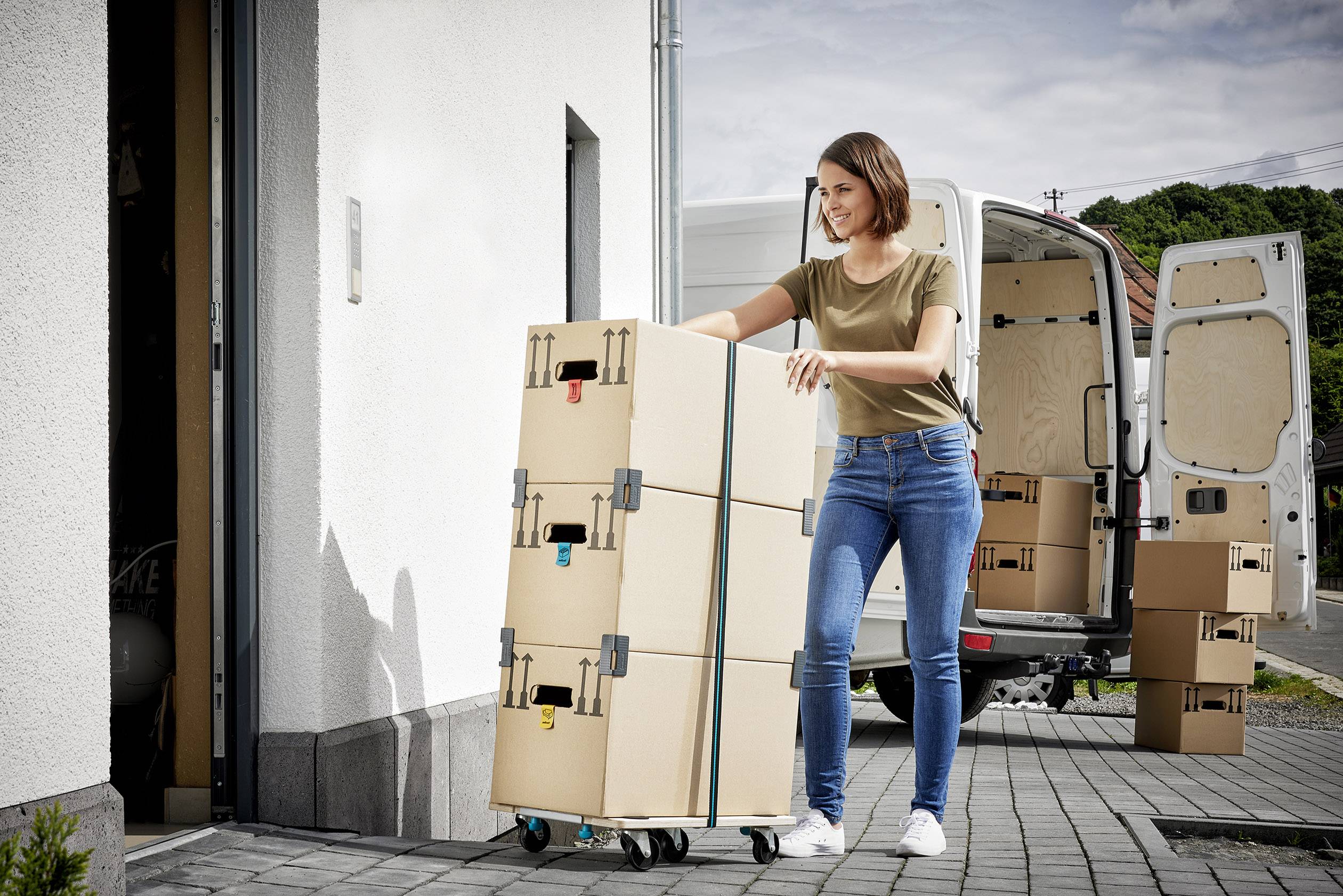 Frau schiebt gestapelte Umzugskartons auf einem Rollwagen in ein Gebäude. Ein Lieferwagen mit geöffneten Türen steht im Hintergrund.