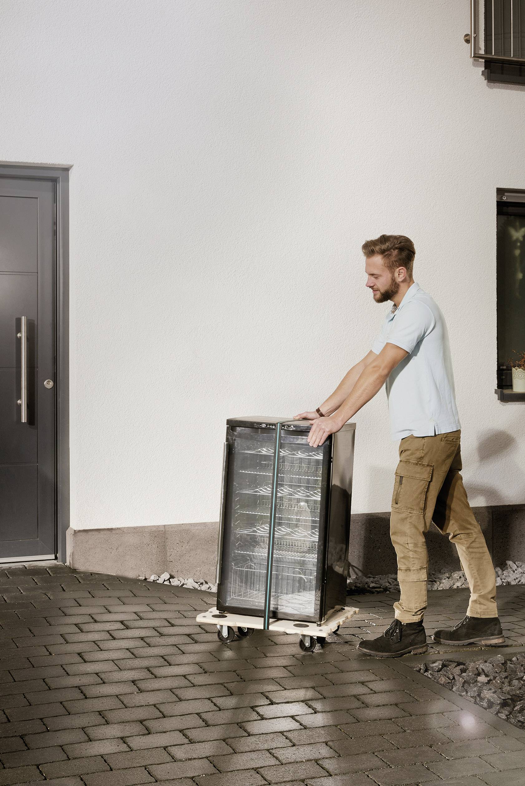 Eine Person schiebt einen schwarzen Kühlschrank auf einem Rollwagen über eine gepflasterte Einfahrt in der Nähe eines Hauses.