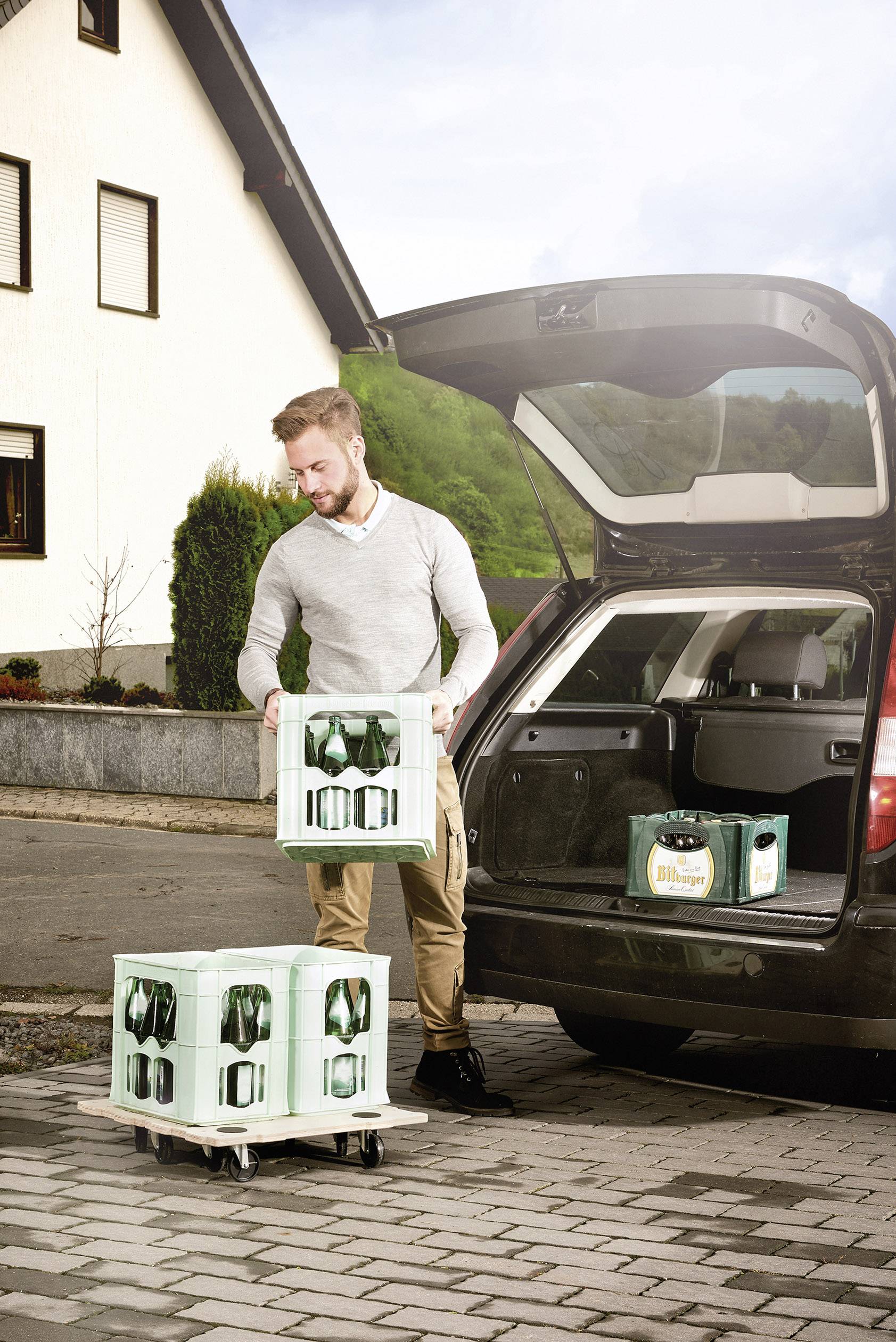 Ein Mann lädt Getränkekisten aus dem Kofferraum eines Autos auf einem Parkplatz aus. Im Hintergrund steht ein Wohnhaus.