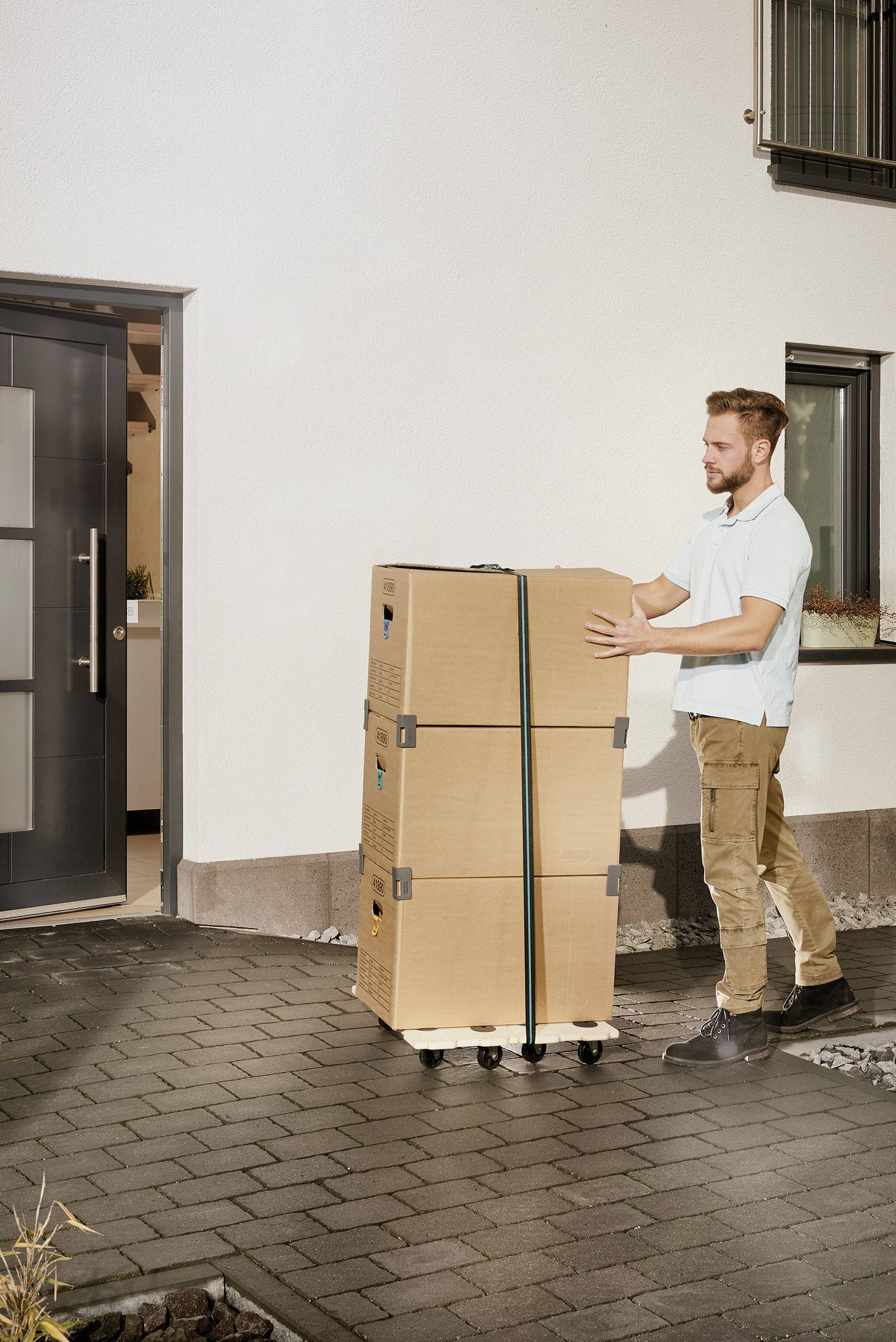 Ein Mann transportiert drei gestapelte Kartons auf einem Rollbrett vor einem modernen Hauseingang.