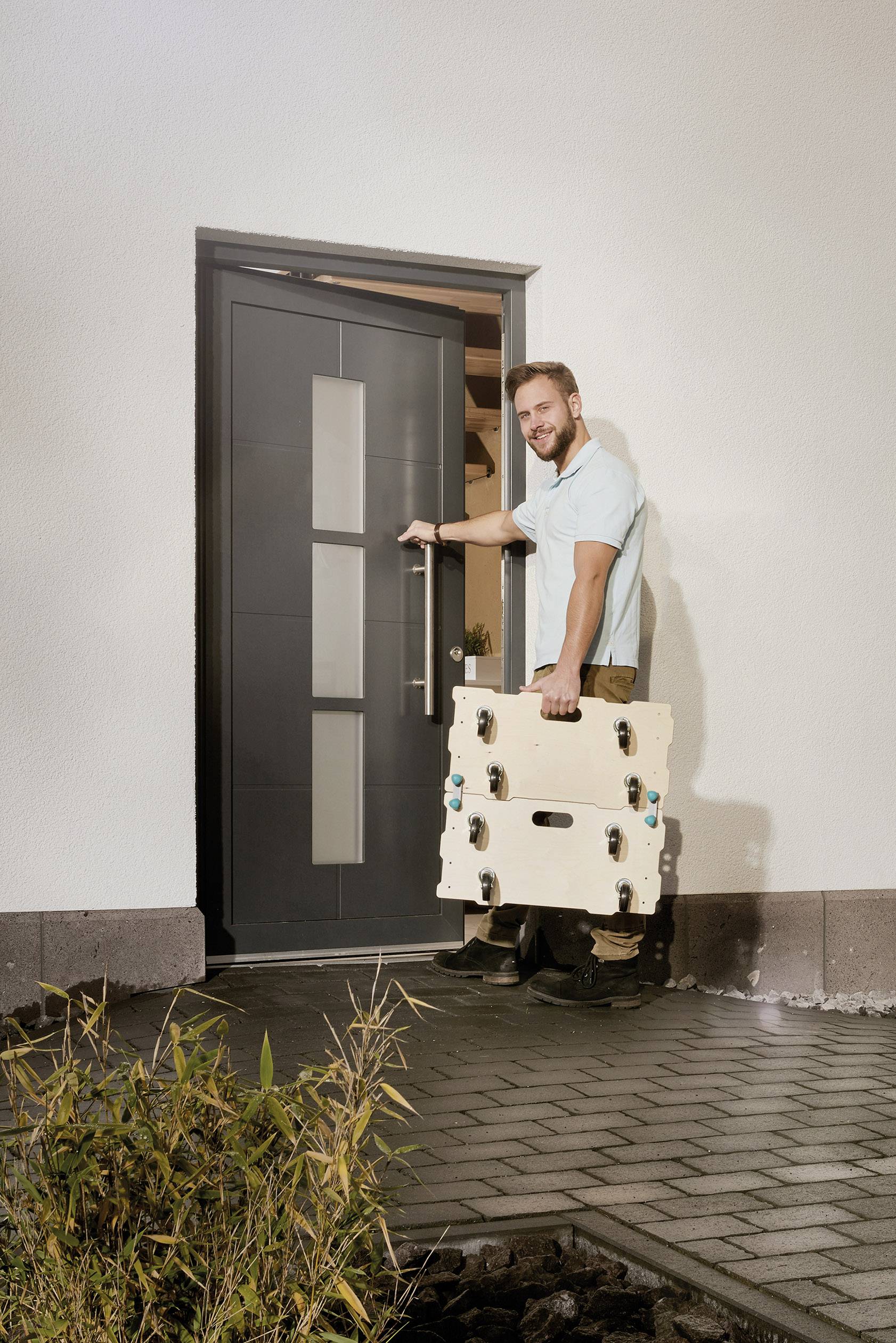 Ein Mann öffnet eine Haustür und hält einen großen, beige Koffer mit Rollen, bereit hineinzutreten. Im Vordergrund sind Pflanzen sichtbar.