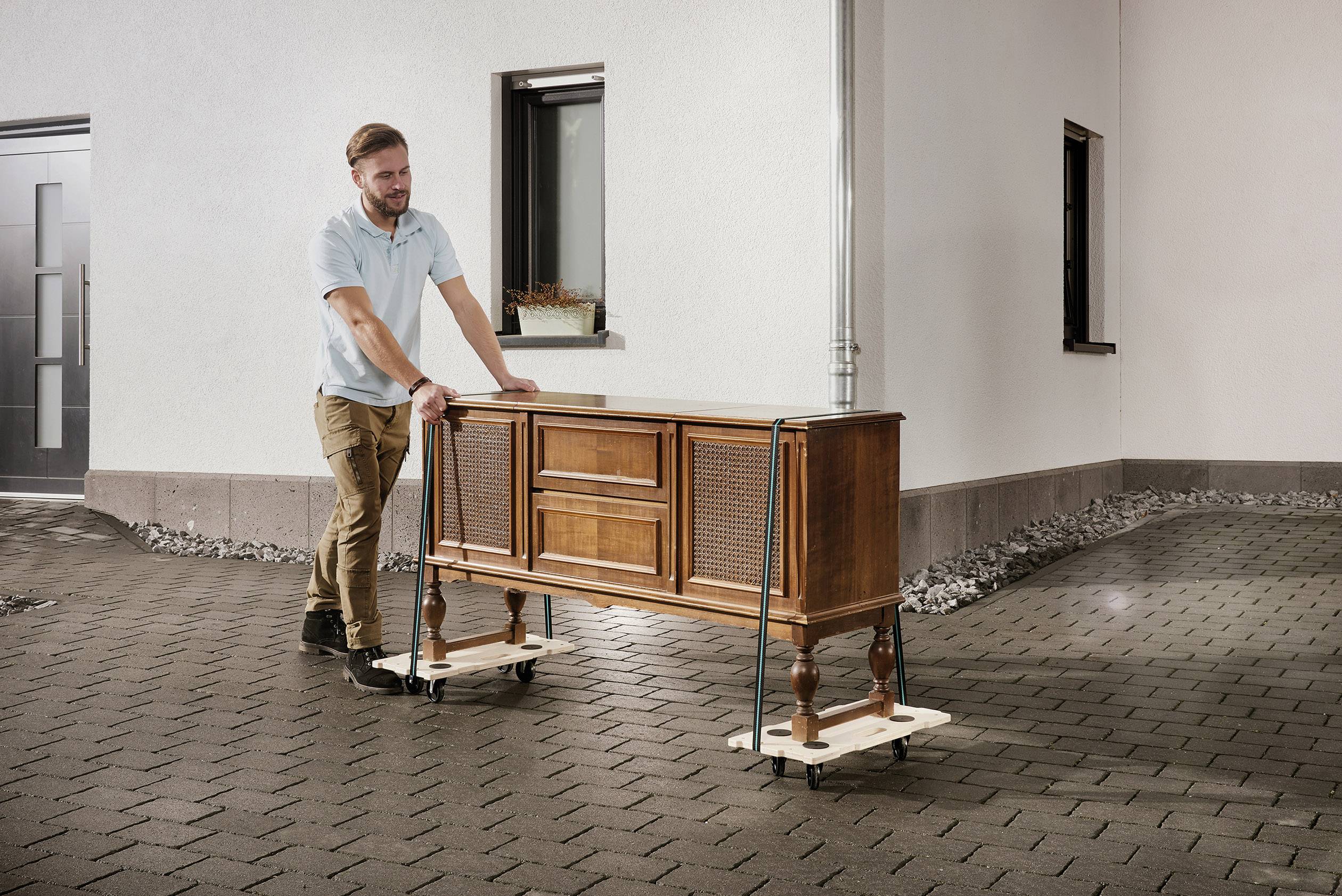 Eine Person bewegt ein antikes Möbelstück auf Rollbrettern über einen gepflasterten Hof. Das Möbelstück ist mit Gurten gesichert.