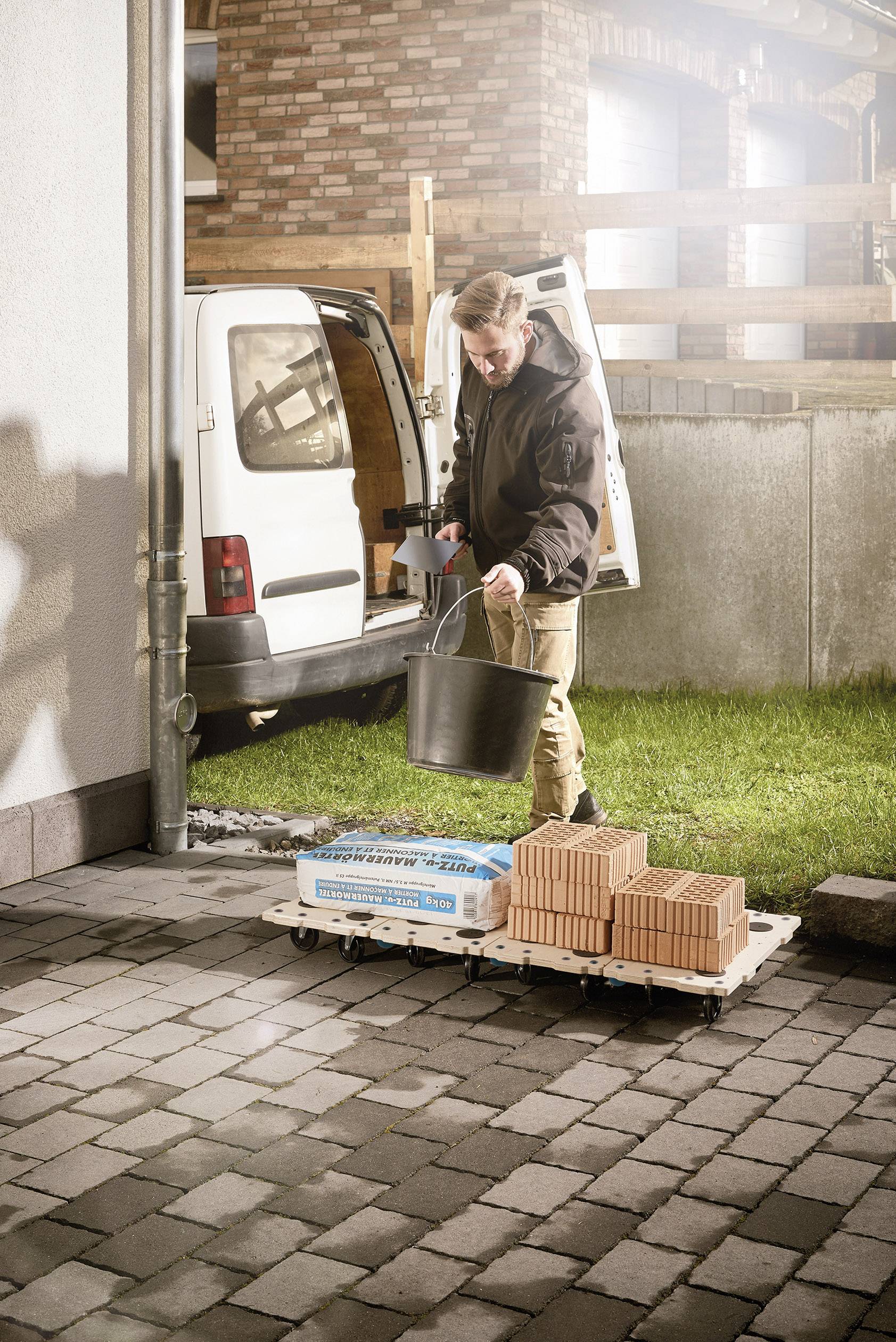 Ein Mann entlädt eine Palette mit Ziegeln und einer Tüte Zement vor einem Van in einem Hof. Er trägt einen Eimer.