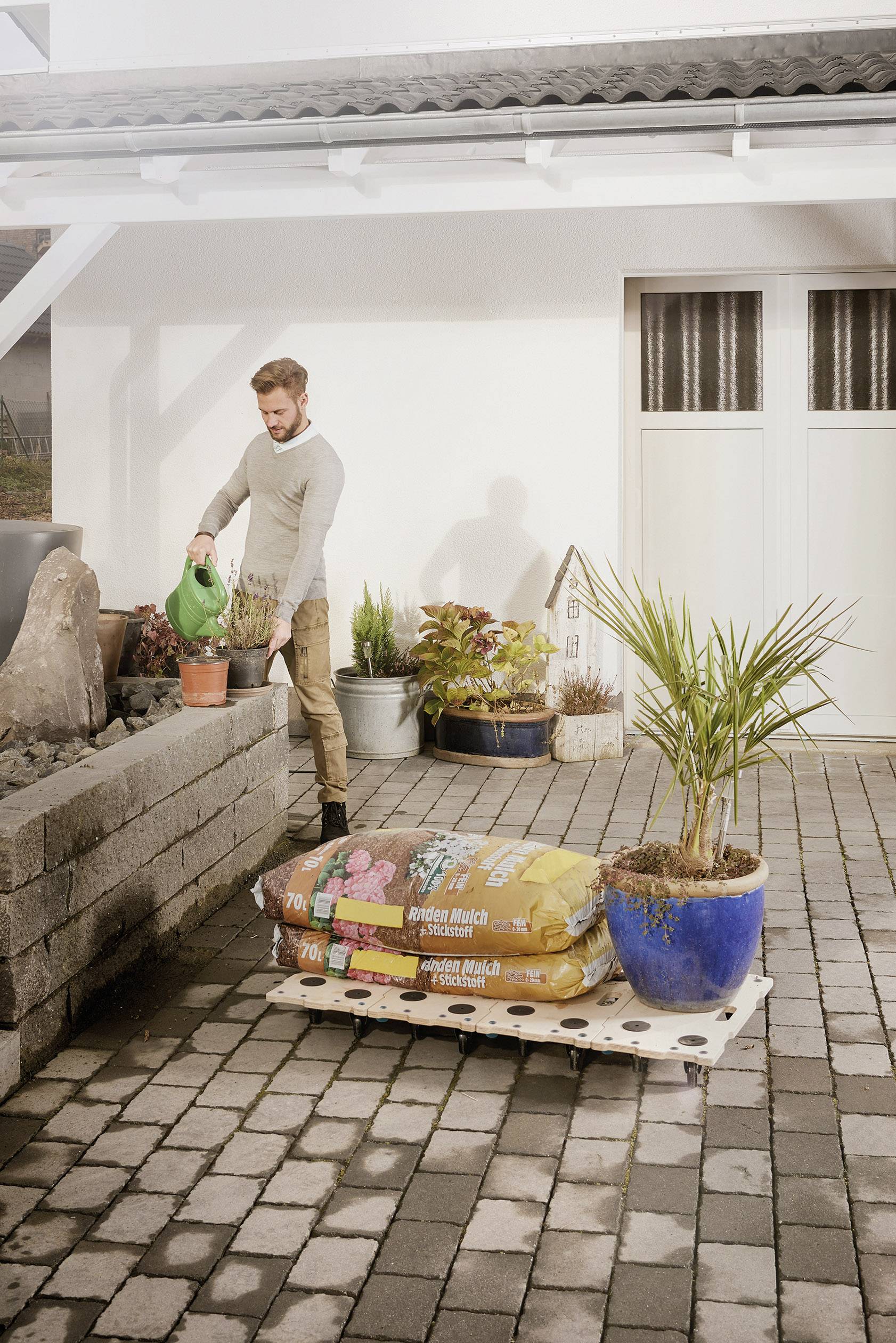 Ein Mann gießt Pflanzen in einem Garten mit Pflastersteinen. Im Vordergrund stehen Pflanzsäcke und ein großer Blumentopf.
