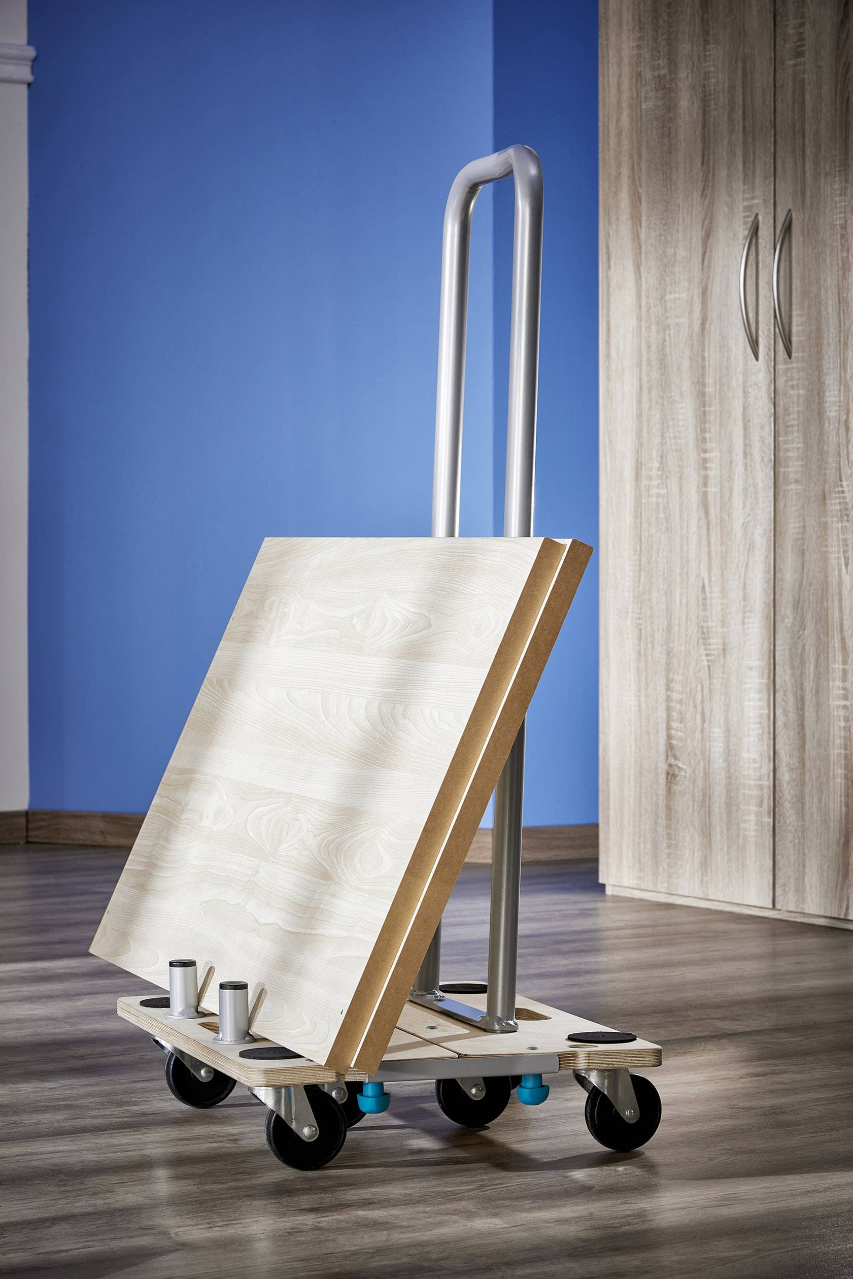 Ein Transportwagen mit zwei Holzplatten steht auf einem Holzboden. Im Hintergrund sind ein blauer Wandabschnitt und ein Holzschrank zu sehen.