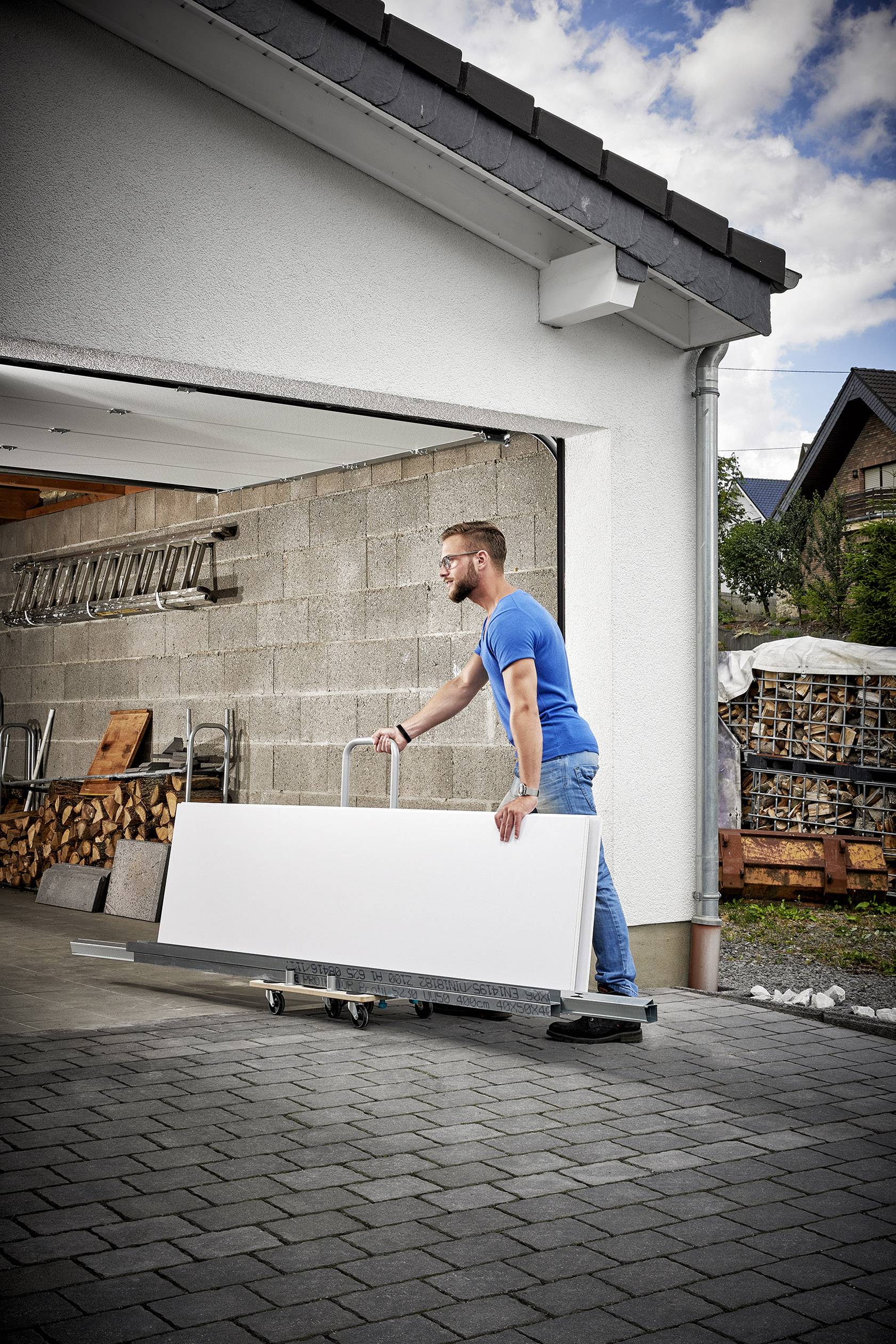 Mann in blauem T-Shirt bewegt ein großes, weißes Panel auf einem Rollwagen in eine Garage. Im Hintergrund Holzstapel und Leiter.