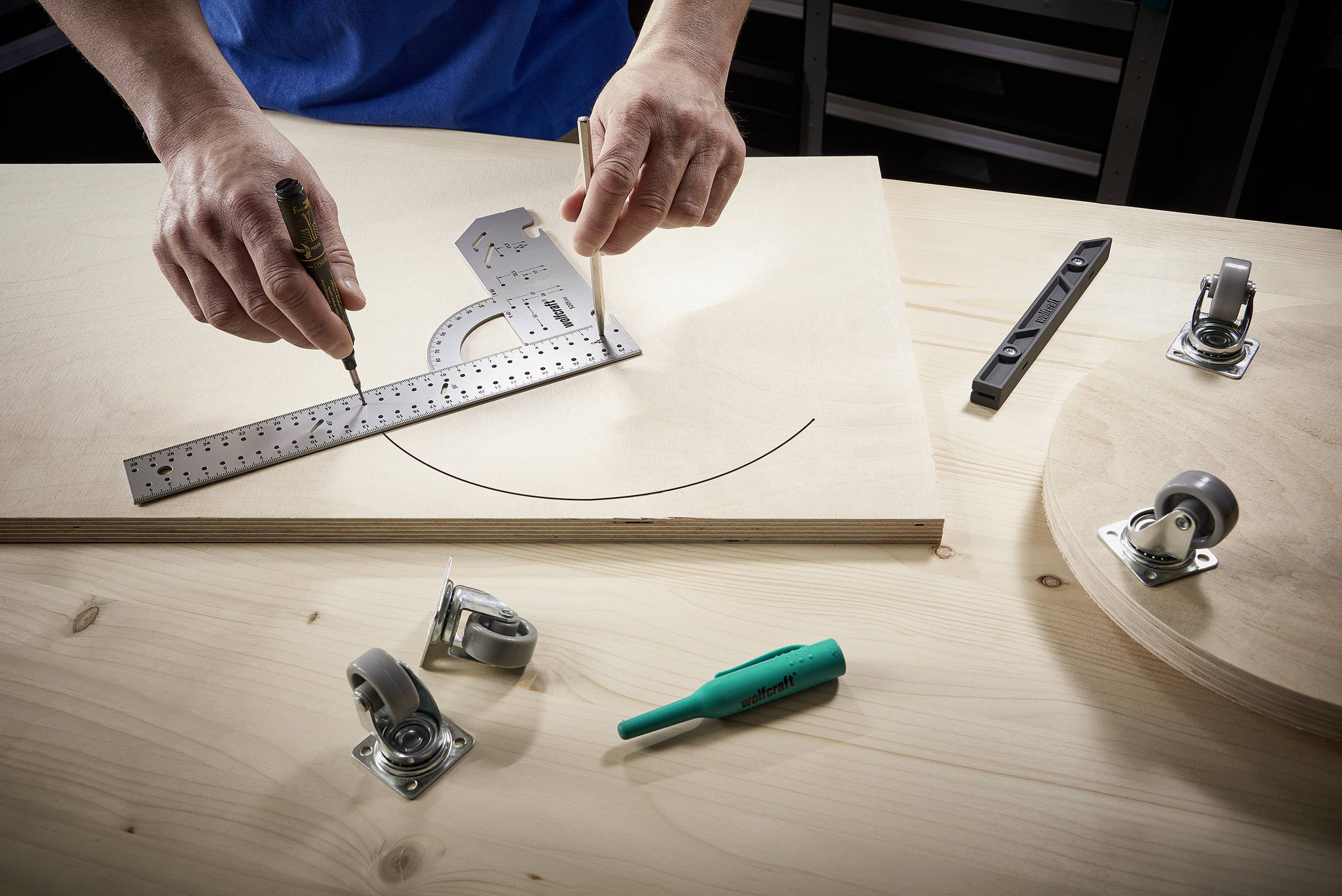 Eine Person zeichnet mit einem Stahllineal und einem Zirkel einen Bogen auf eine Holzplatte. Auf dem Tisch liegen Räder und ein Marker.
