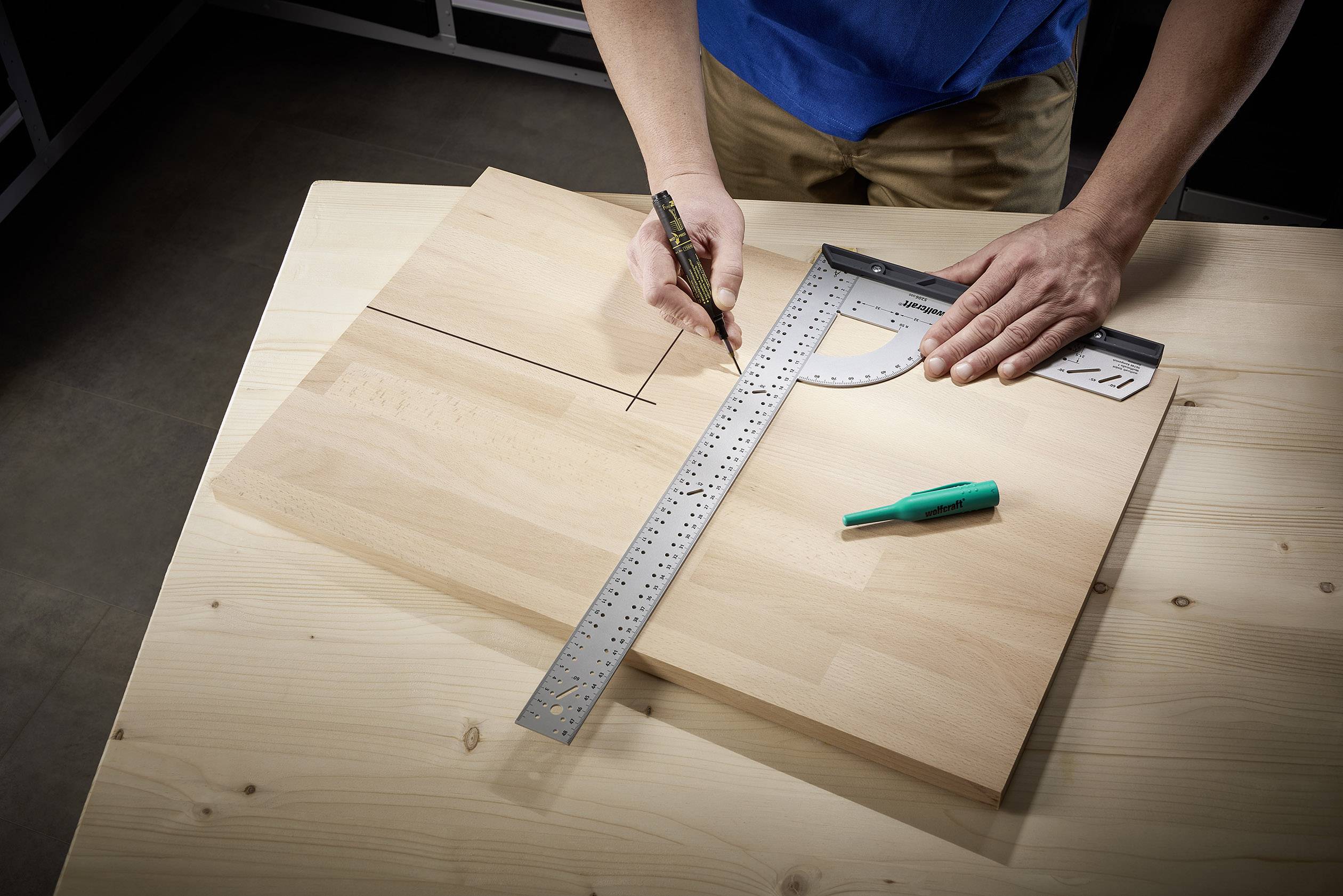 Person misst auf einem Holztisch ein Holzbrett mit einem Geodreieck und Bleistift aus. Ein grüner Stift liegt daneben.
