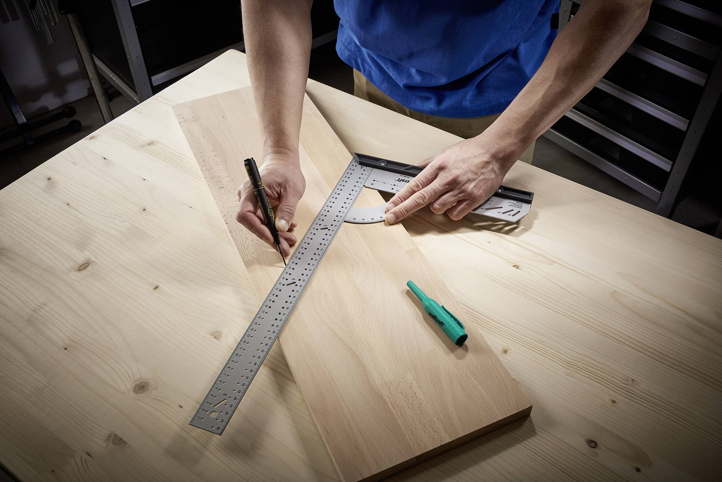 Eine Person zeichnet mit einem Winkelmesser und Lineal Linien auf ein Holzbrett. Ein grüner Stift liegt daneben auf dem Tisch.