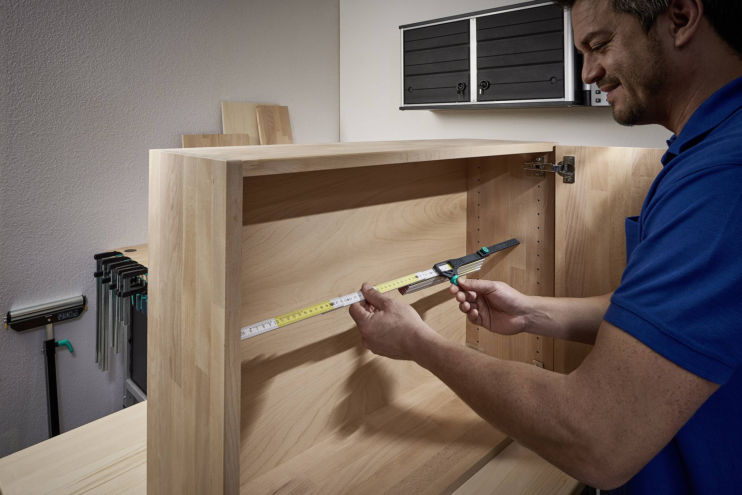 Ein Mann misst die Innenbreite eines Holzschranks mit einem Maßband in einer Werkstatt. Werkzeuge und Holzbretter sind im Hintergrund.