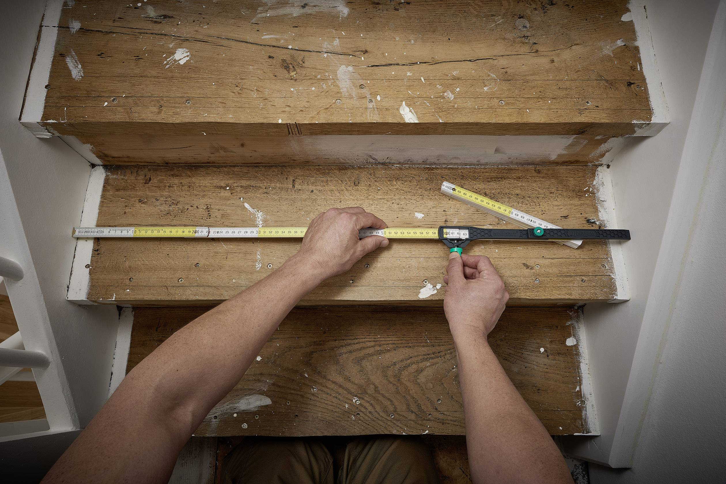 Eine Person misst die Breite und Tiefe einer hölzernen Treppenstufe mit zwei Maßbändern, die auf der Stufe platziert sind.