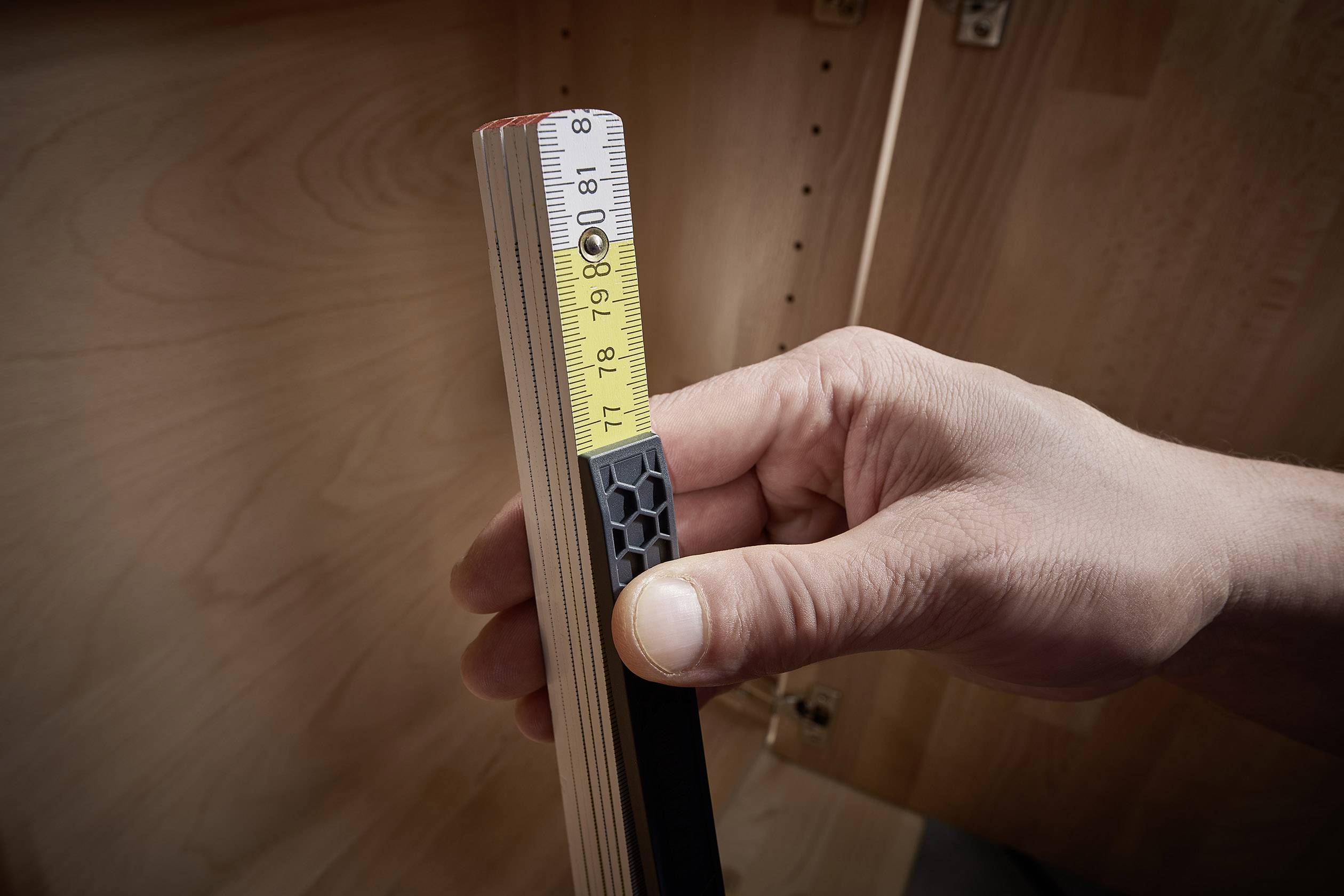 Eine Hand hält ein ausziehbares Holzmaßband, das eine Länge von etwa 9 Zoll zeigt. Im Hintergrund sind Holzmöbel sichtbar.