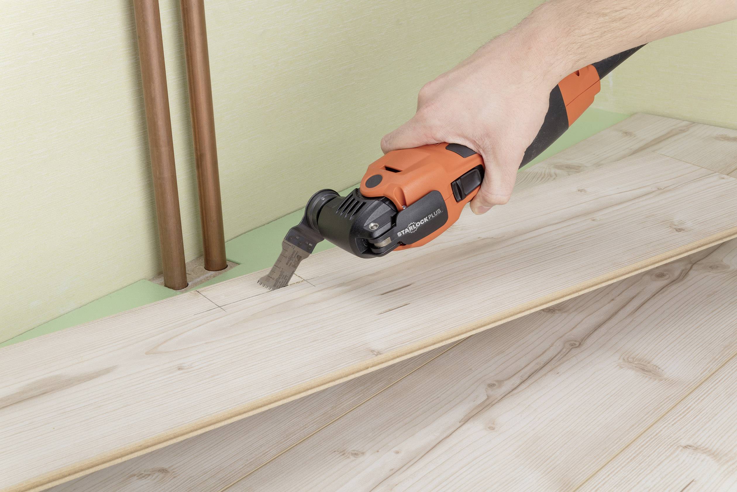 Eine Person schneidet mit einem Multitool ein Stück Holz, das Teil einer Bodenverlegung ist. Copperrohre sind im Hintergrund zu sehen.