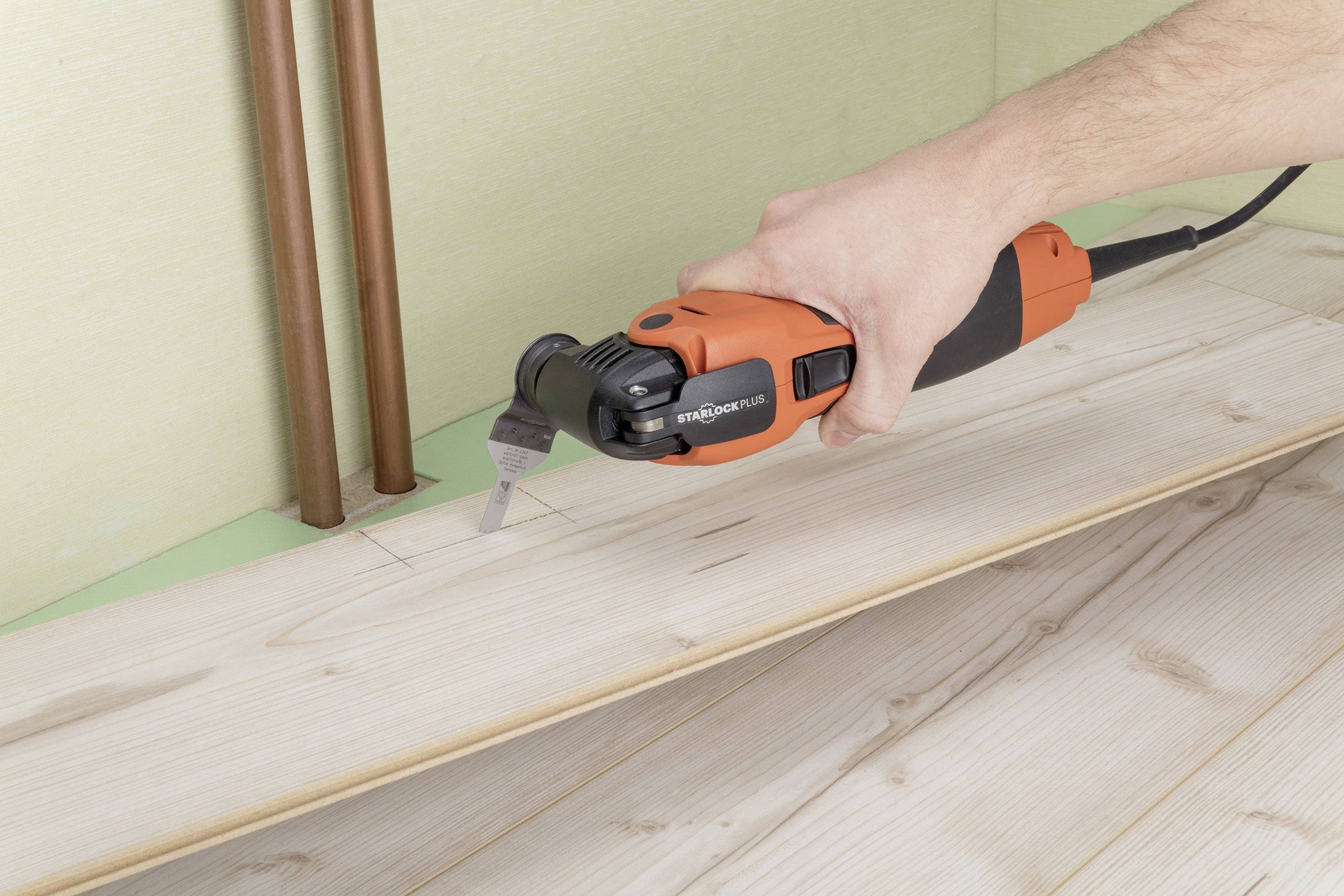 Eine Person schneidet mit einem oszillierenden Multifunktionswerkzeug eine Aussparung in ein Holzbodenbrett in der Nähe von Rohren.