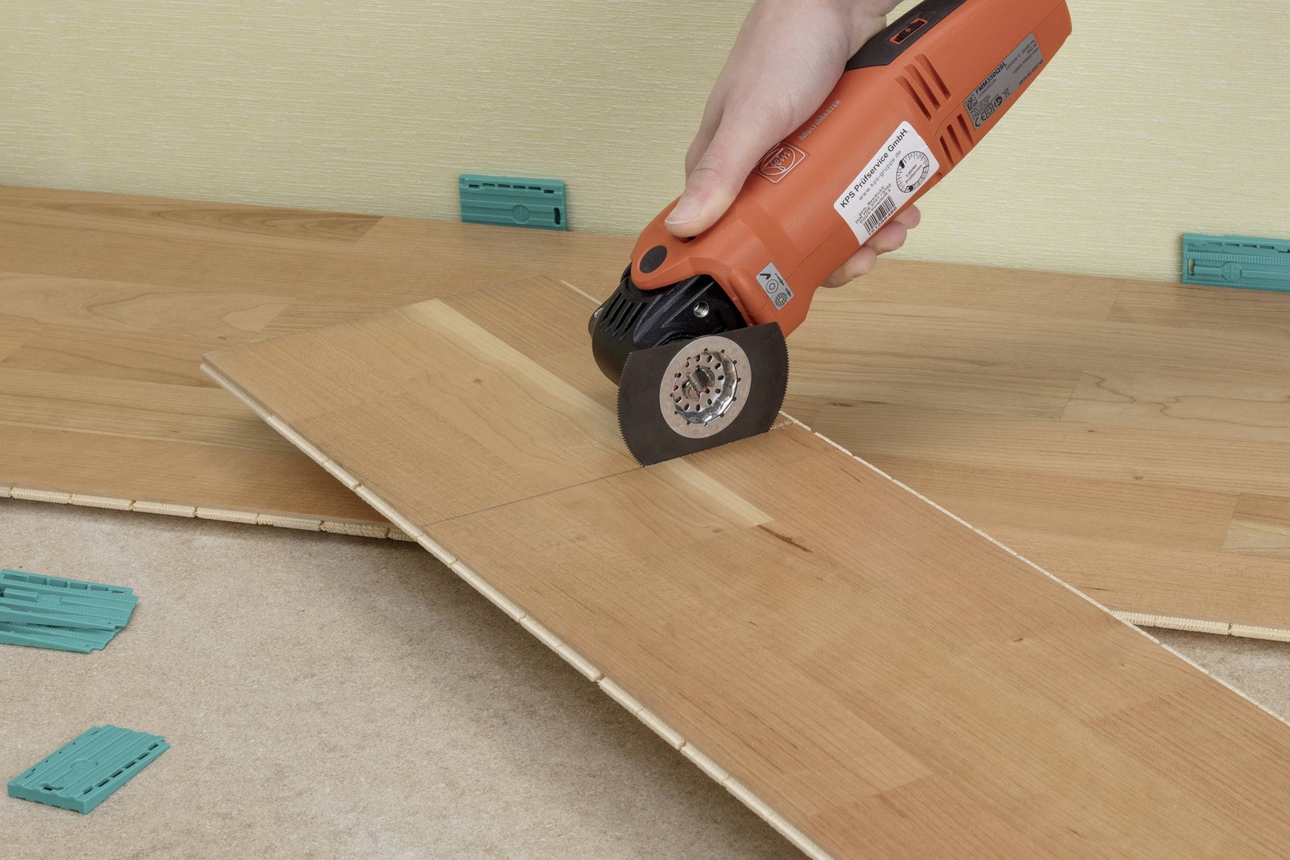 Eine Hand hält ein oszillierendes Multifunktionswerkzeug, das eine Holzplatte schneidet. Einige Abstandshalter liegen auf dem Boden.