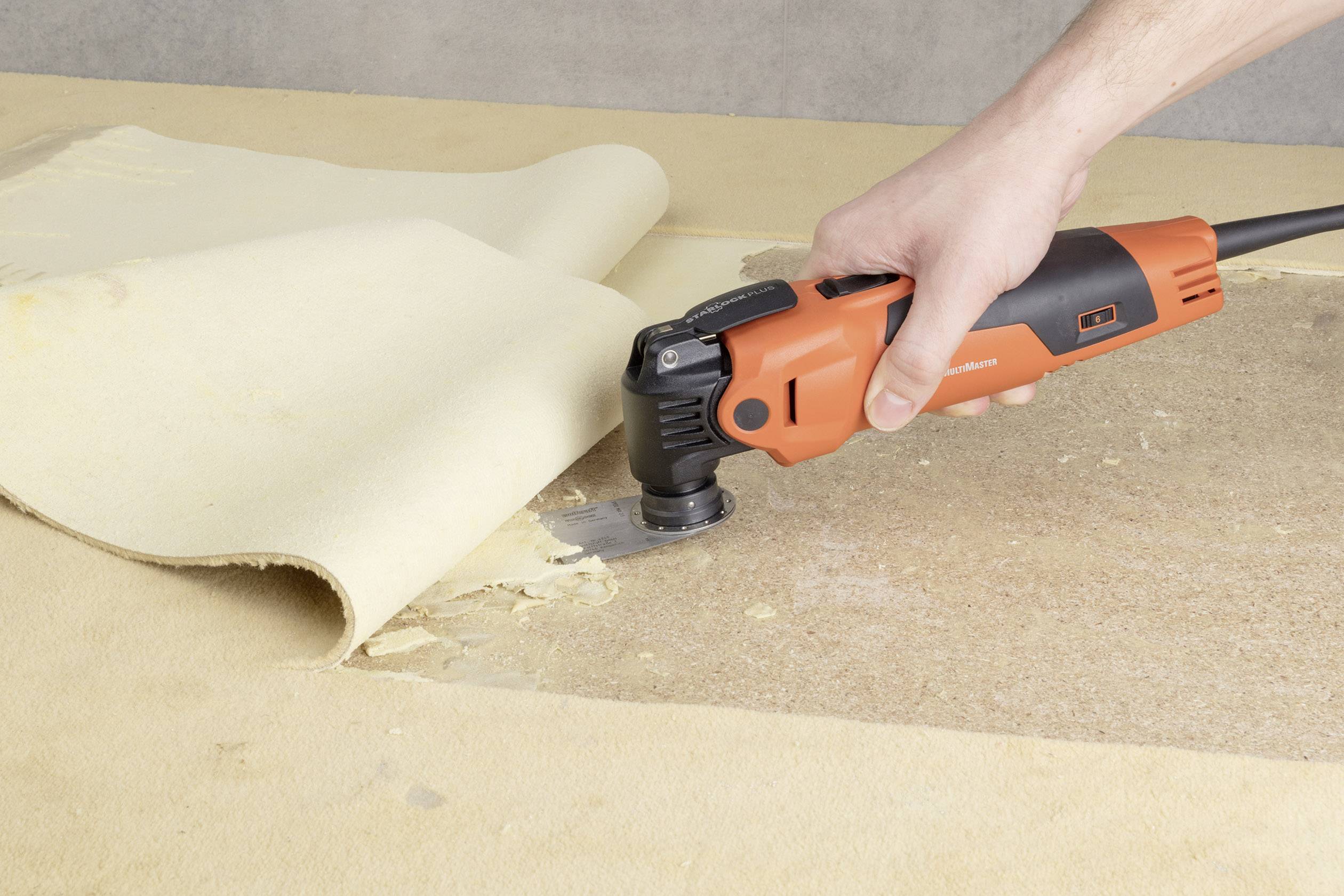 Eine Person verwendet ein orangefarbenes Elektrowerkzeug, um Teppich von einem Holzboden zu entfernen.