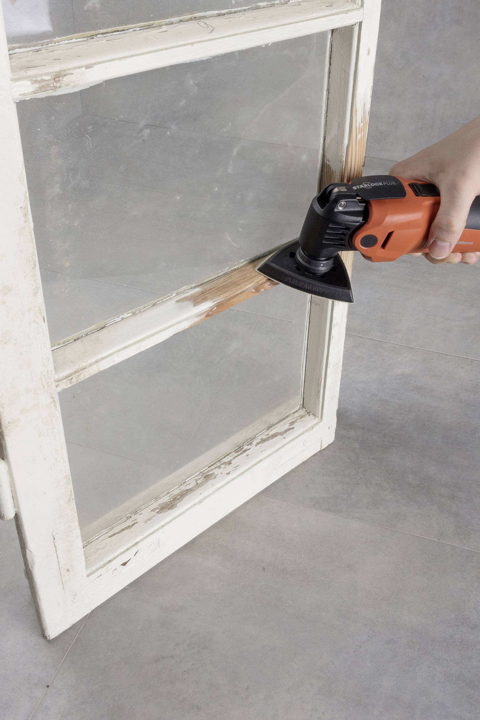 Eine Person schleift alten Lack von einem Holzfensterrahmen mit einem elektrischen Schleifgerät ab. Der Rahmen wird renoviert.