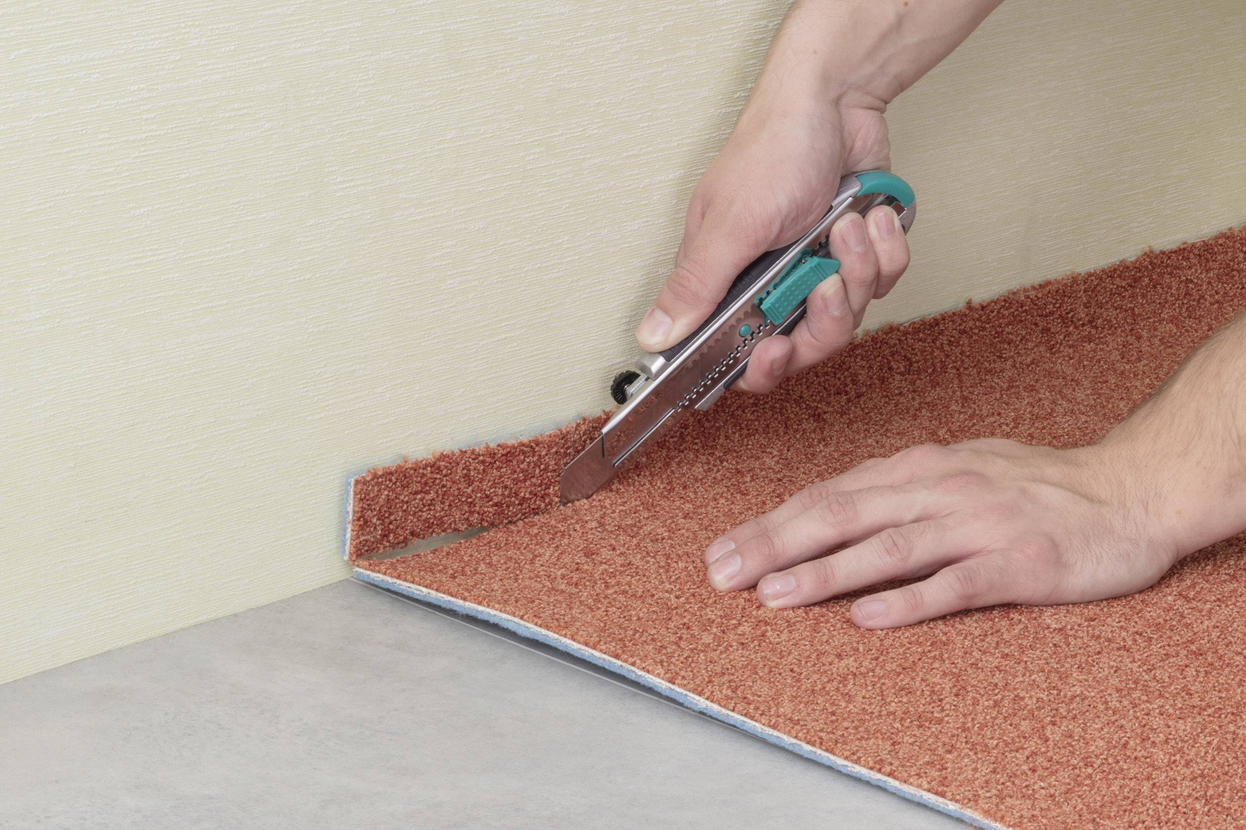 Eine Person schneidet mit einem Cuttermesser einen rotbraunen Teppich entlang einer Kante. Der Teppich liegt auf einem Bodenbelag.
