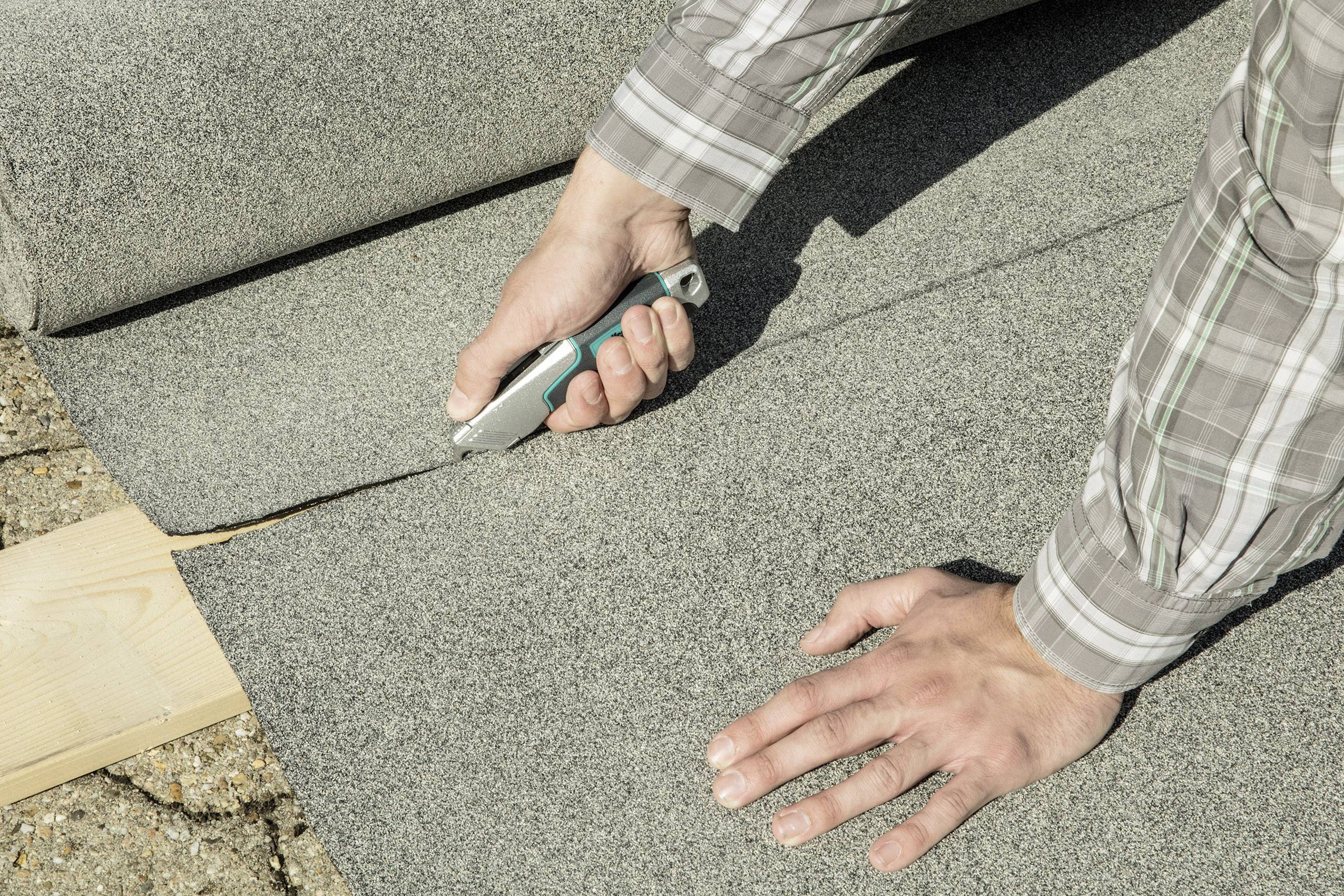 Person schneidet grauen Teppich mit einem Cuttermesser auf einem Holzbrett zurecht. Hände und Teil des Werkzeugs sichtbar.