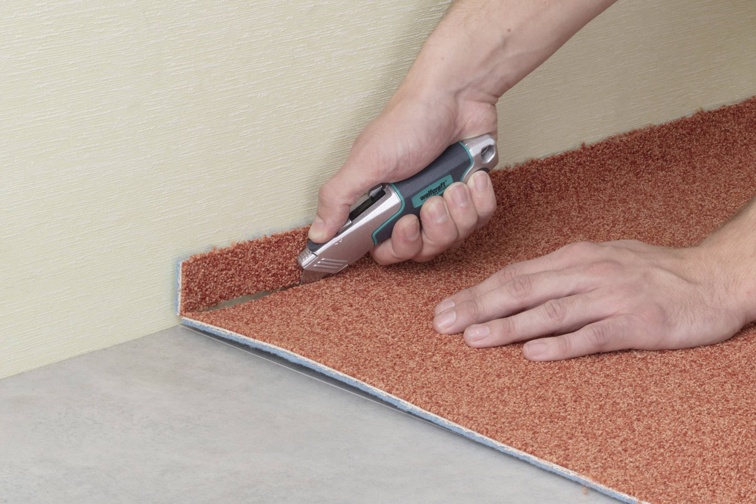 Eine Person schneidet einen orangen Teppich entlang einer Wand mit einem Teppichmesser, um ihn anzupassen.