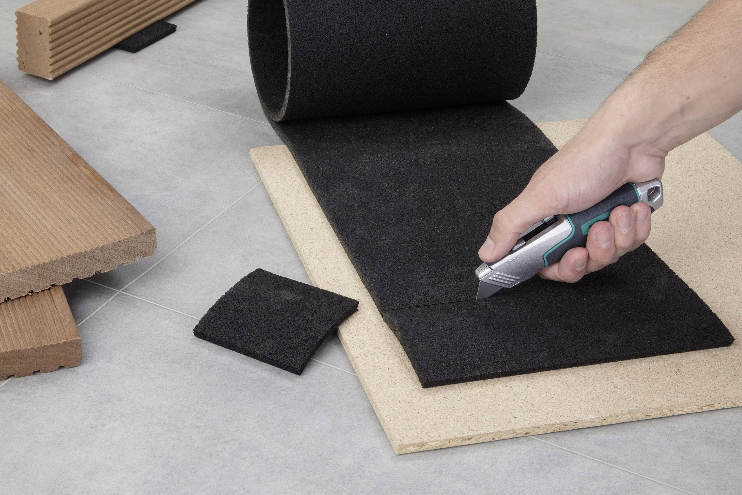 Eine Person schneidet mit einem Cutter-Messer ein Stück schwarzes Belagsmaterial auf einer Holzplatte. Im Hintergrund sind Holzstücke zu sehen.